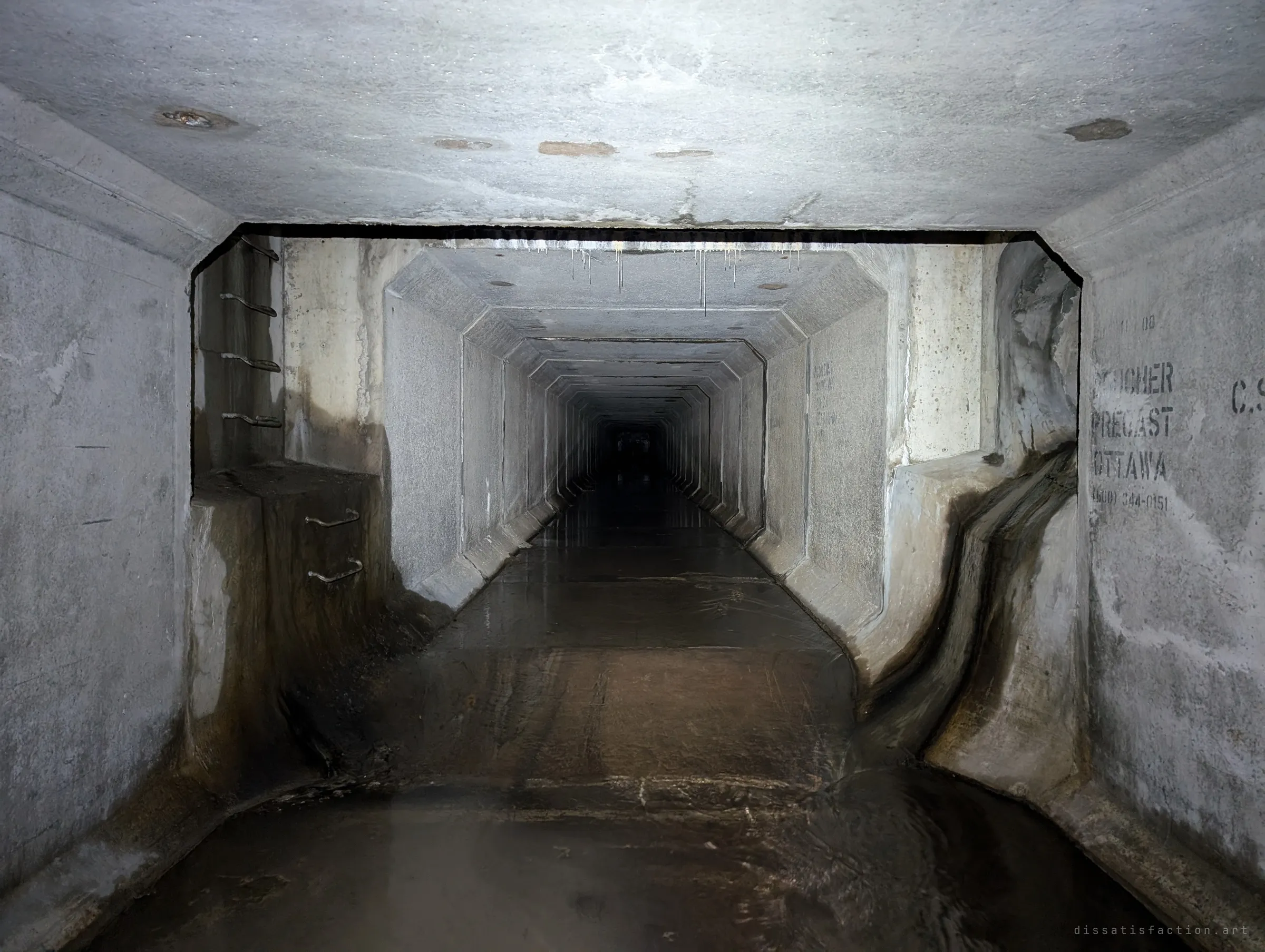 A junction where the culvert steps into a narrower tunnel that runs straight into darkness, with climbing rungs set into the left wall and a dark water stain streaking the right corner. The tunnel seems to go on forever.