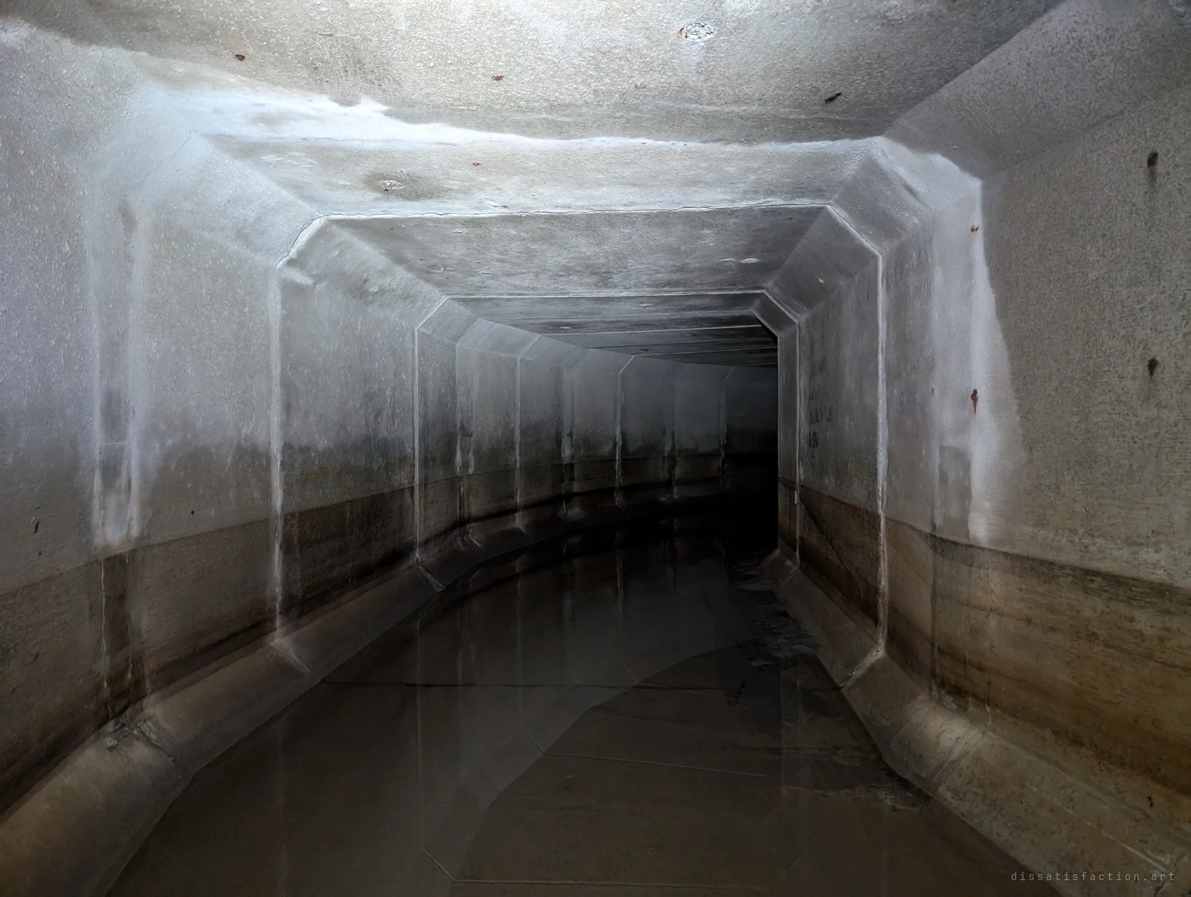 A plain stretch of box culvert curving gently to the right, a brown tide line marking how high the water usually runs. Nothing but concrete, water and the bend ahead.