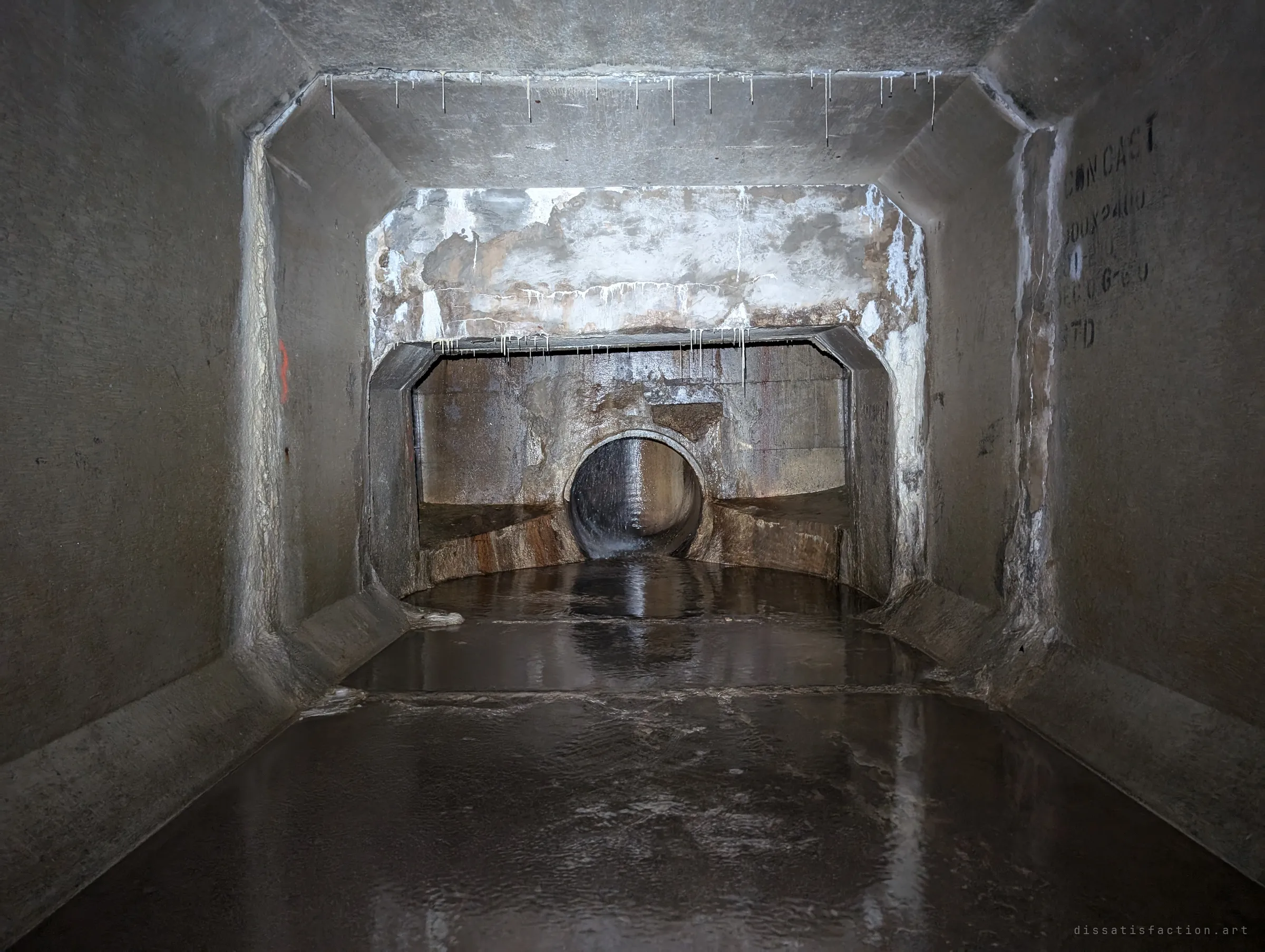 Two culvert sections meet badly, the far one sitting lower and offset, with rough patched concrete above the step and a round pipe pouring into the chamber beyond. A mismatch the builders never quite hid.