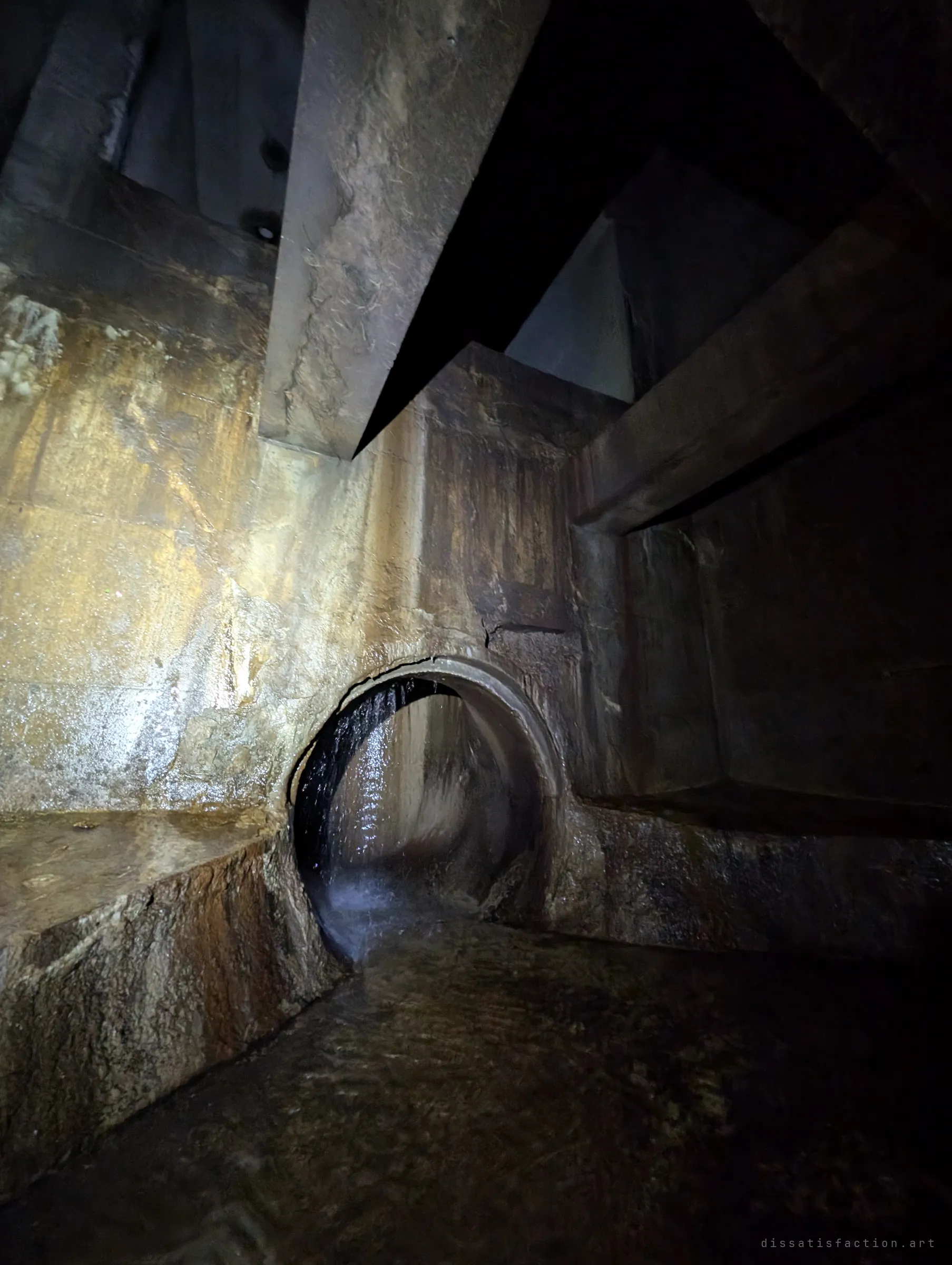 Looking up inside a dropshaft chamber, heavy concrete baffles jut overhead while water spills from a round pipe at its base. The light catches the wet, stained walls of the shaft.
