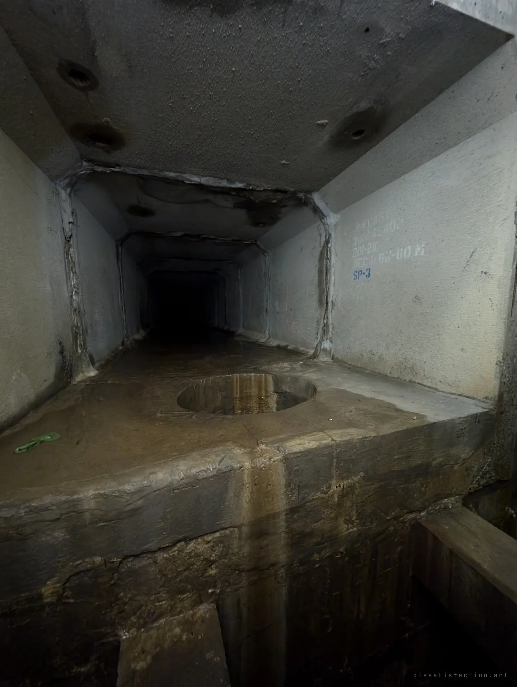 A low precast box culvert stretches into darkness beyond a raised concrete ledge with a round drop hole cut into it. Stencilled product codes on the wall mark the segment type.