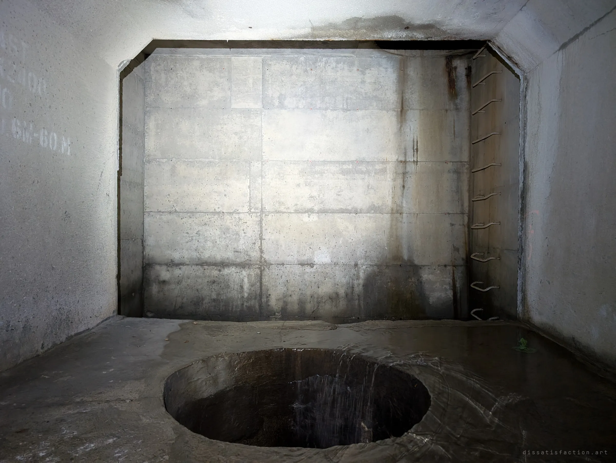 A round drop shaft opens in the wet floor of a concrete chamber, with a line of bent steel rungs climbing the far corner. Water trickles over the lip into the dark below.
