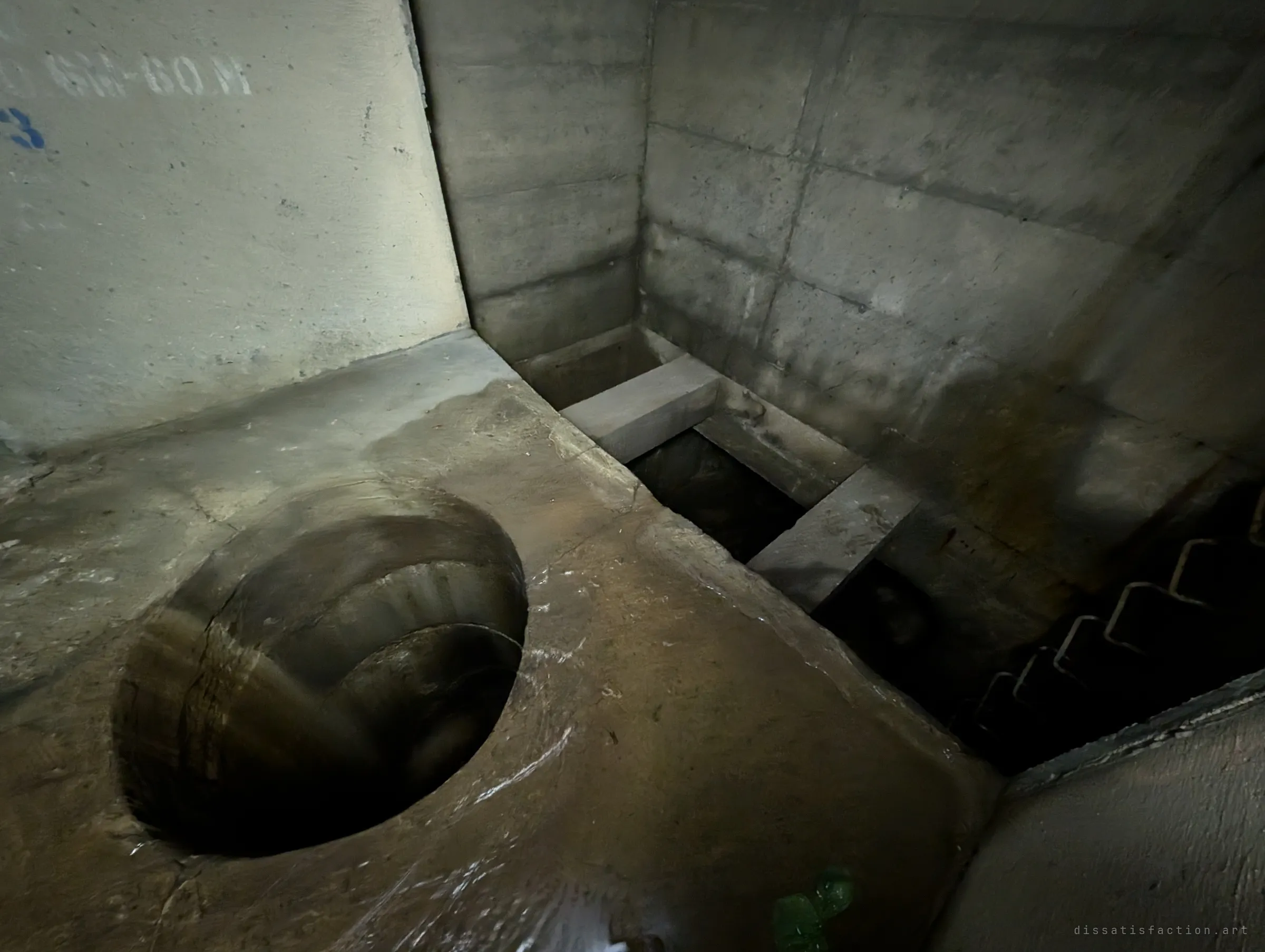 Looking down beside the drop hole into a lower channel crossed by two concrete beams, with rungs descending the side wall. It shows how the chamber steps down between levels.
