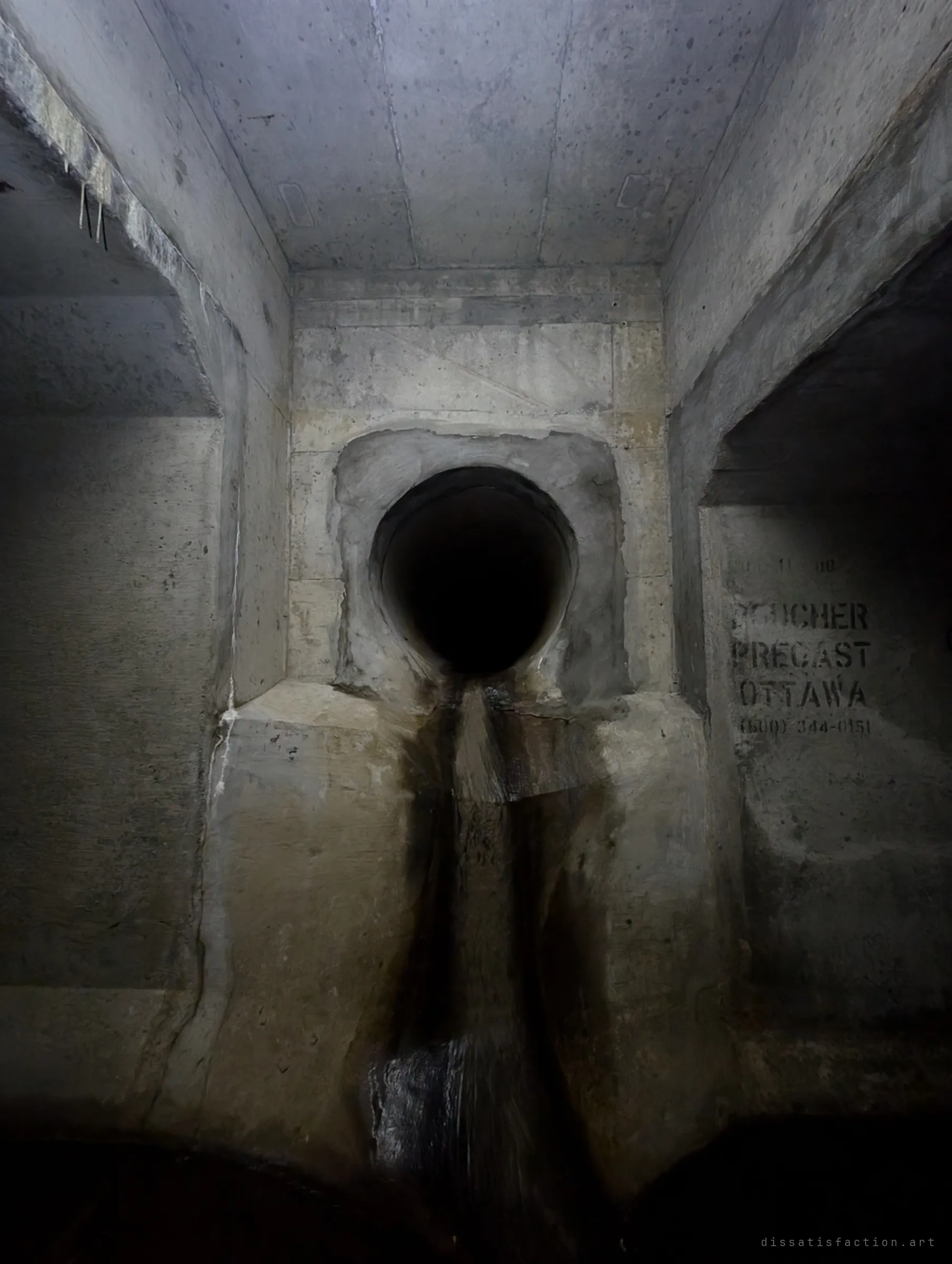 A dark round pipe outlet sits in the end wall of a concrete junction, a dark stain of runoff streaking down beneath it. Side openings lead off to the left and right.