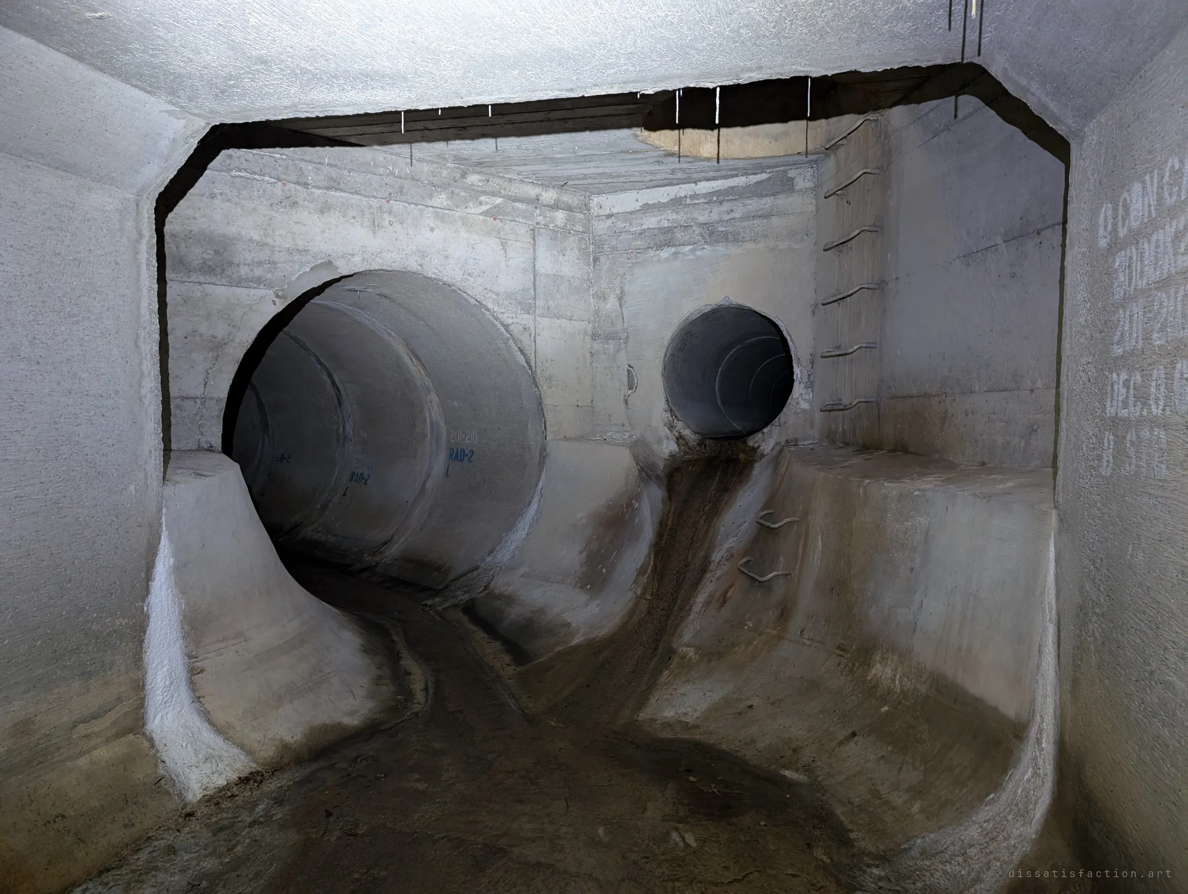 Two round pipes of different sizes empty into a shaped concrete junction, with rungs climbing to an access shaft above. The curved benching guides the flows together.