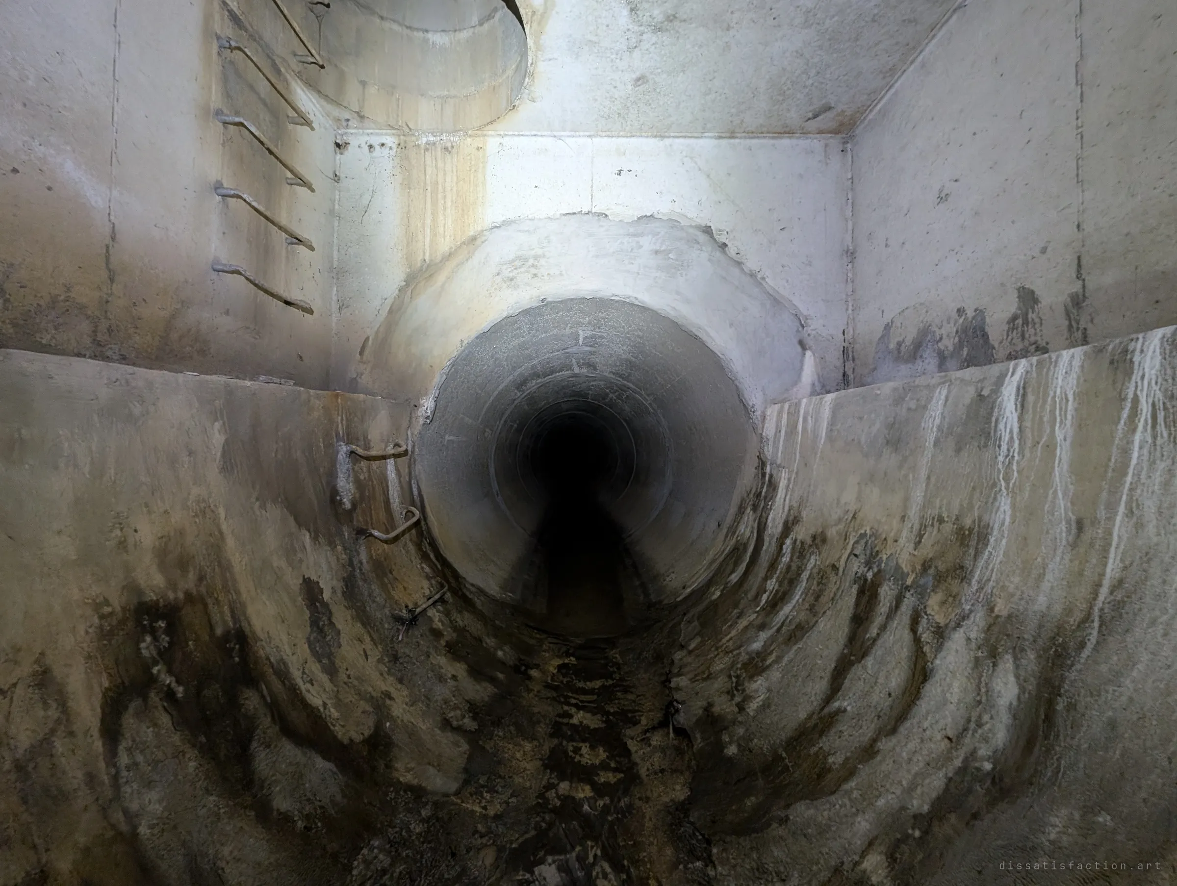 A round pipe tunnels away from a curved concrete trough streaked with mineral runoff, while rungs lead up to a manhole shaft in the ceiling.