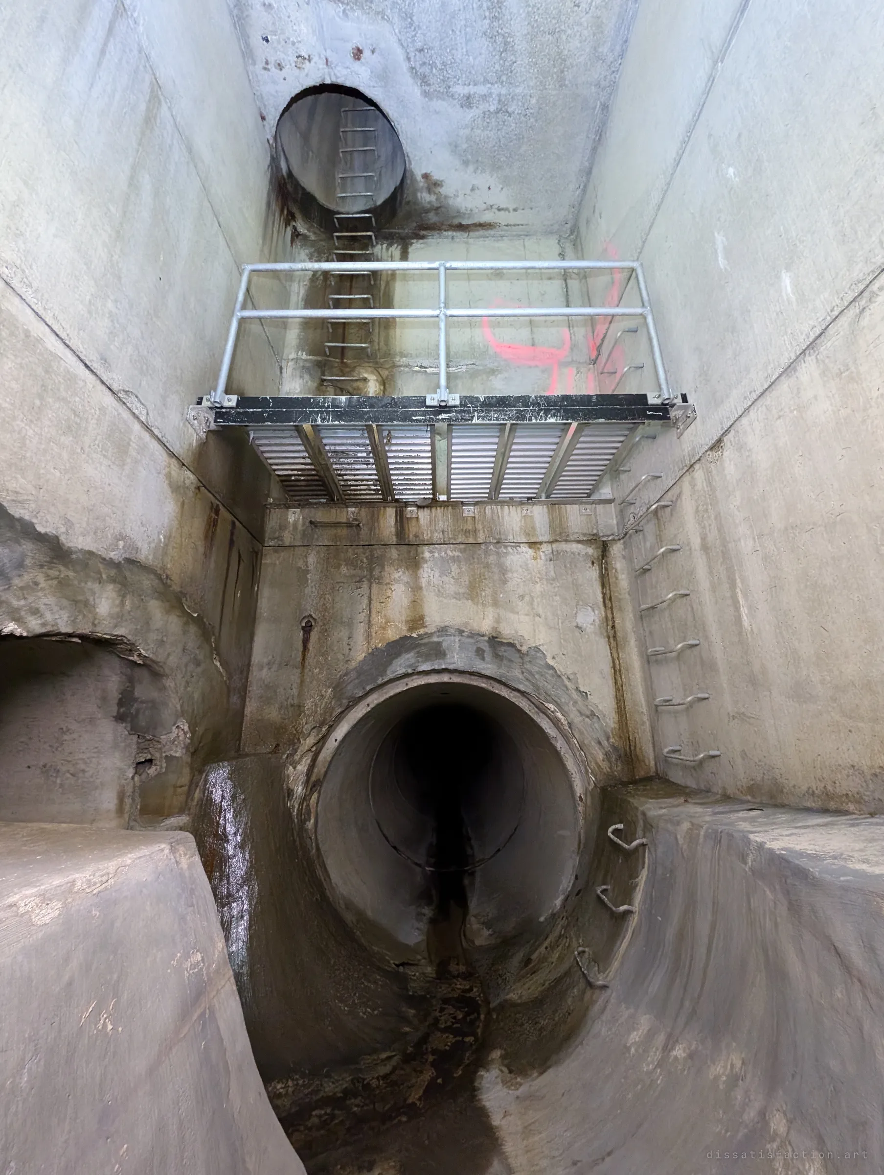 A tall shaft rises above a pipe outlet, with a railed metal landing halfway up and a ladder leading to a round opening at the top. Pink survey paint marks the back wall.