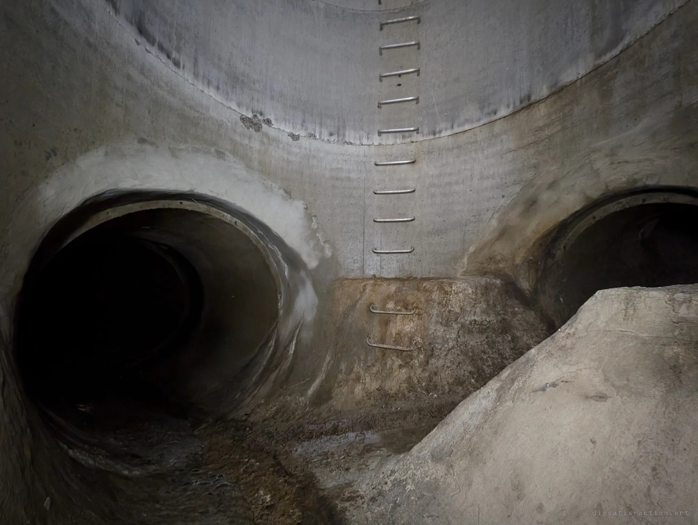 The bottom of a concrete drain shaft, where a line of steel rungs runs down the wall between two dark round pipe outlets and a sloped concrete benching. It shows how the shaft meets the tunnels below.