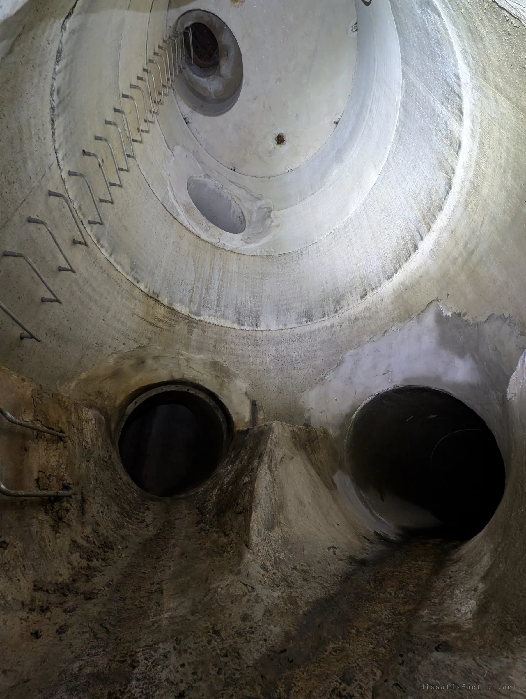 A tilted view up the shaft from its floor, with rungs spiralling along the curved wall towards the grated top and two pipe mouths at the base. The stepped rings of the shaft read like a giant eye.