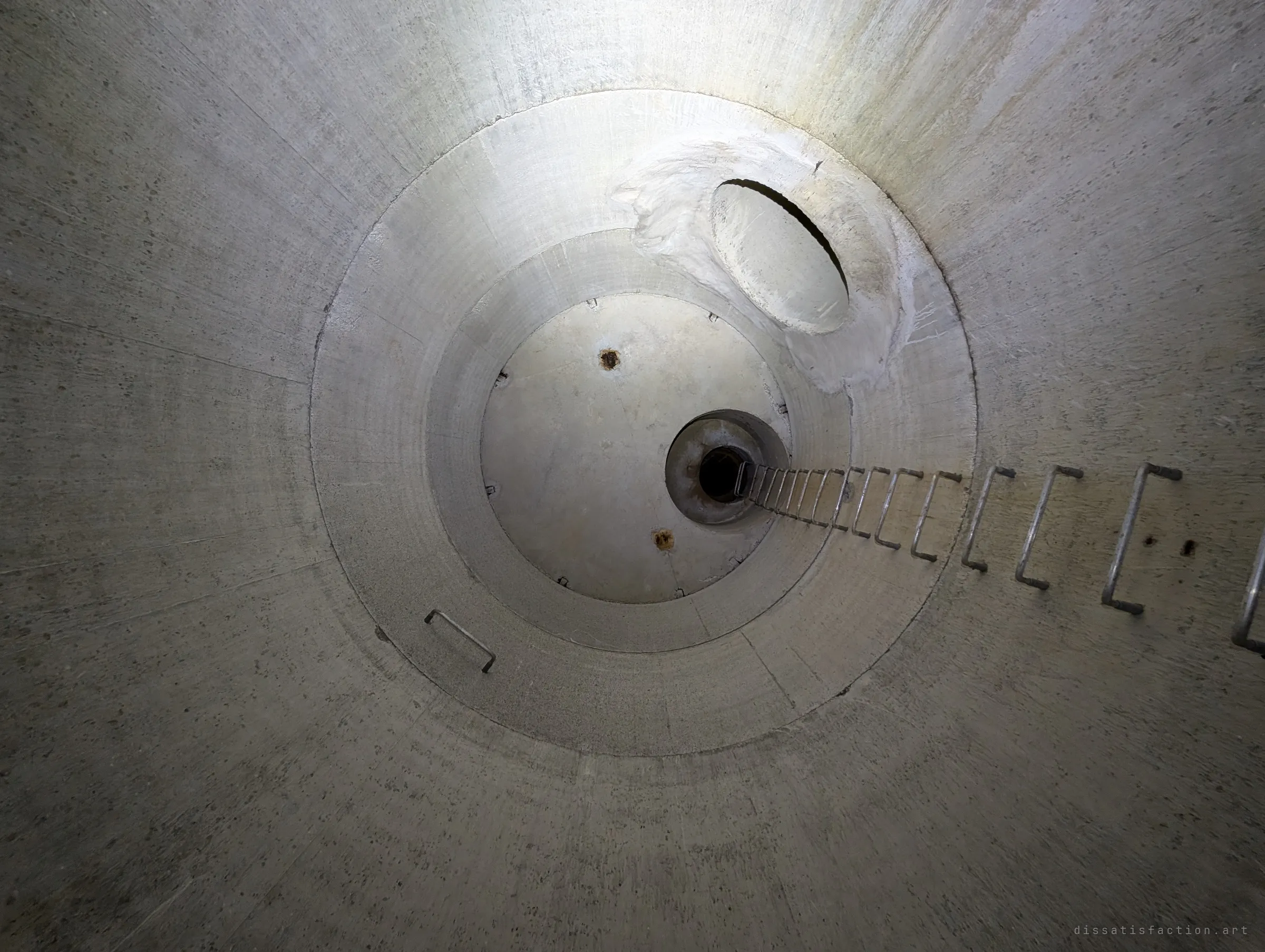 Straight up the shaft: concentric concrete rings narrow to a small dark opening, with a line of rungs leading into it. It shows how far the shaft rises above.