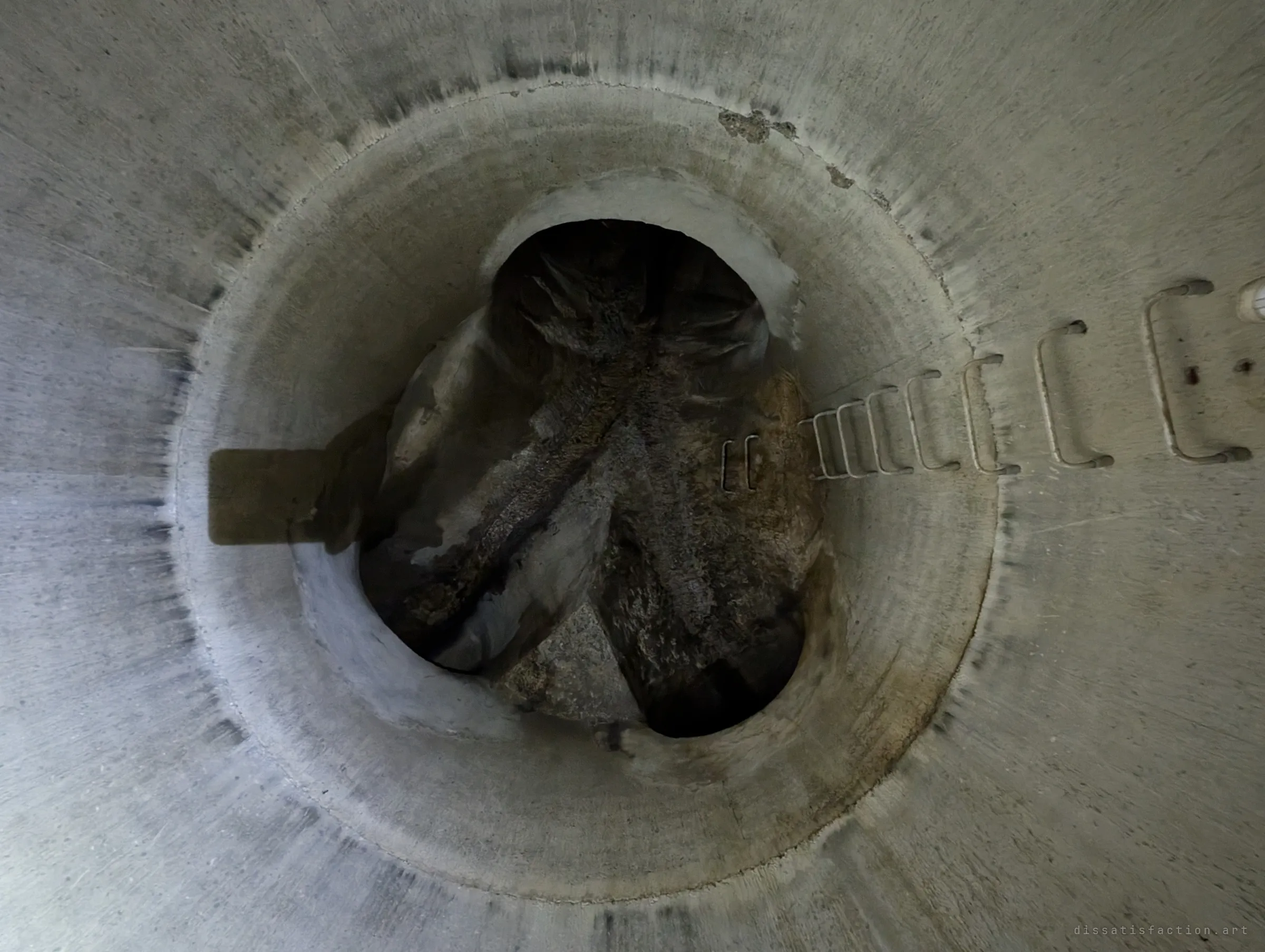 Looking down the shaft from partway up, the rungs lead towards a dim floor where pipe outlets and sloped benching meet. The drop makes the climb feel exposed.