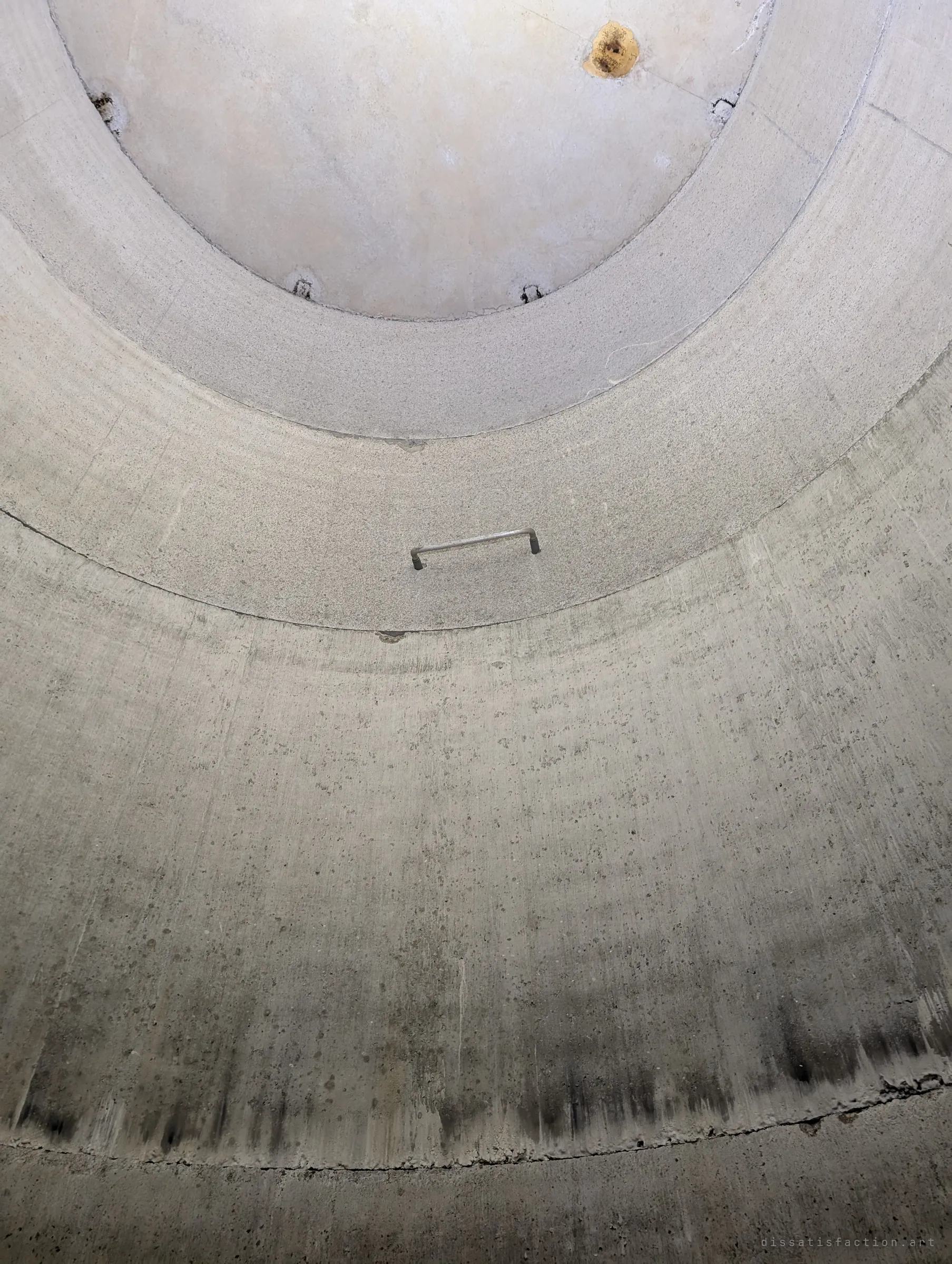 A single steel rung set alone in a bare curved concrete wall, far from any other rung, under the rings of the shaft top. It is one of the lonely rungs that gave the album its name.