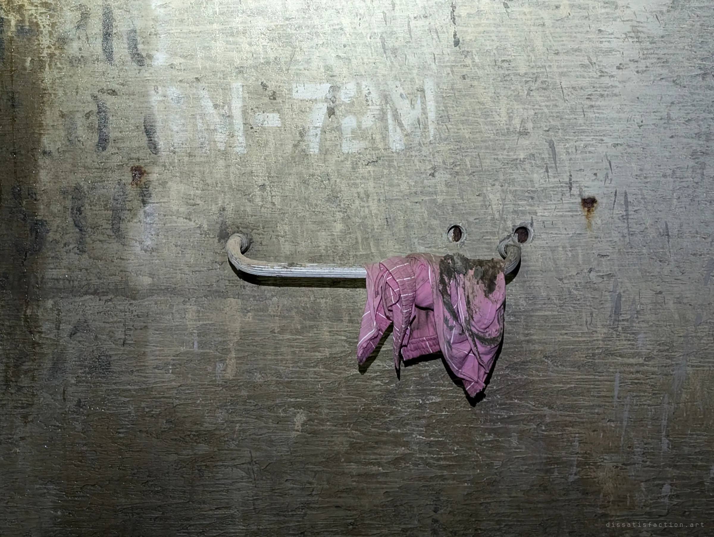 A muddy pink striped cloth hangs from a steel rung on a scarred concrete wall with faded stencilling. A small trace of whoever came through before.