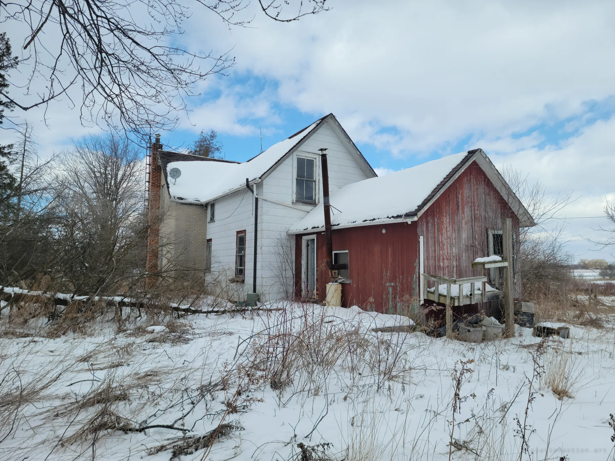 A snowed-in farmhouse seen from the back: white siding, a brick chimney and a weathered red lean-to with a sagging wooden porch. Overgrown brush has crept right up to the walls.