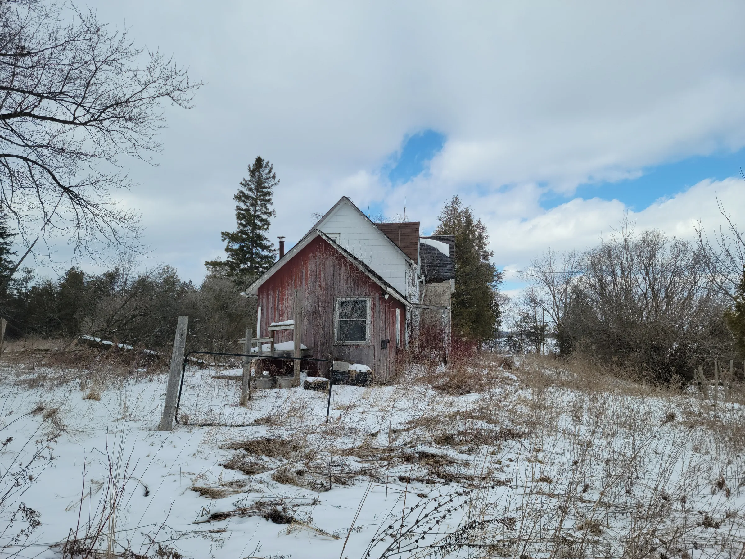 The red back addition of the farmhouse half hidden behind a toppled wire gate and dead grass in the snow. Evergreens and bare trees close in around it.