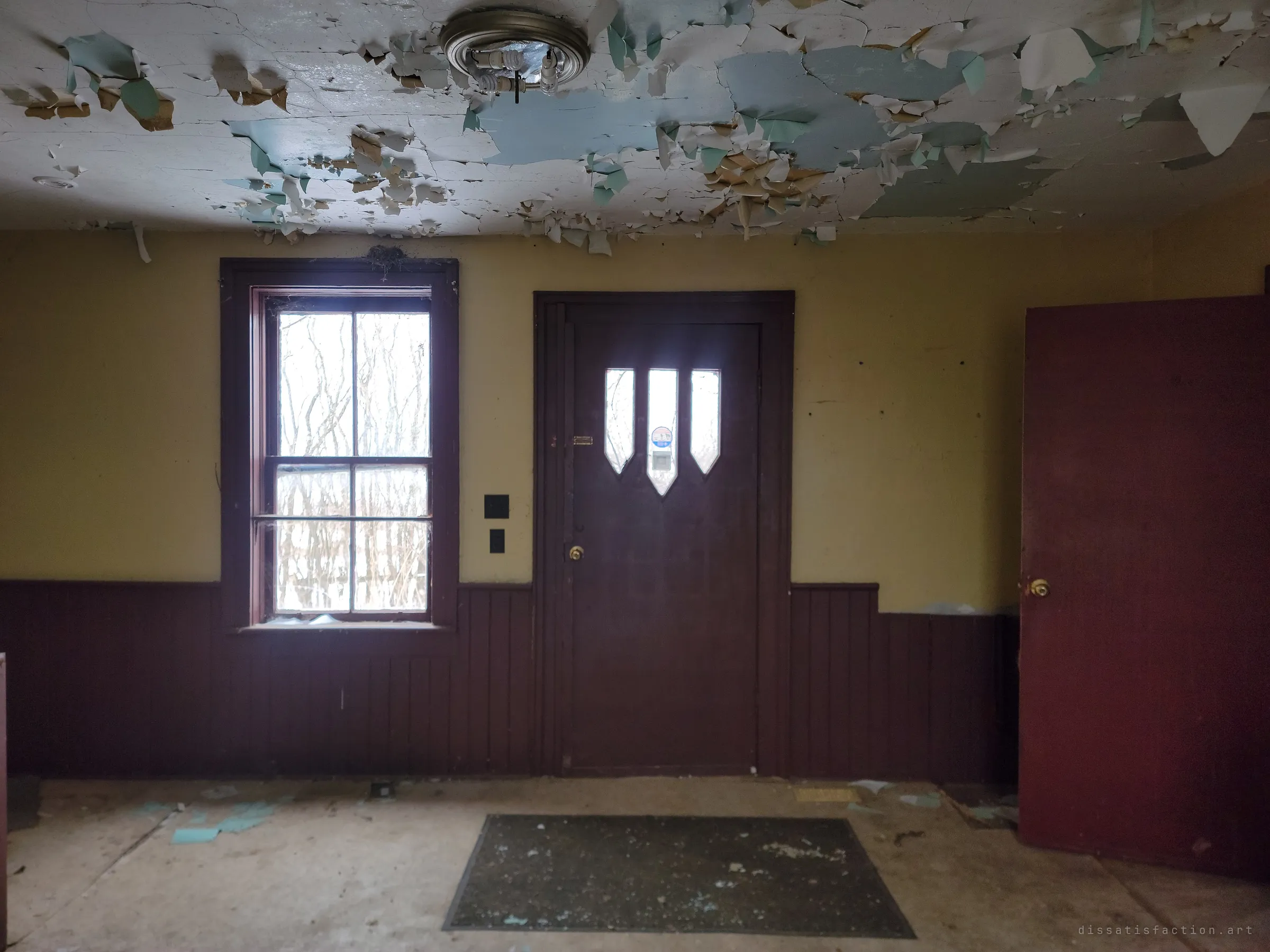 An empty front room with a paint-shedding ceiling, mustard walls and dark wainscoting around a door with three pointed windows. A worn mat still sits where people once wiped their boots.