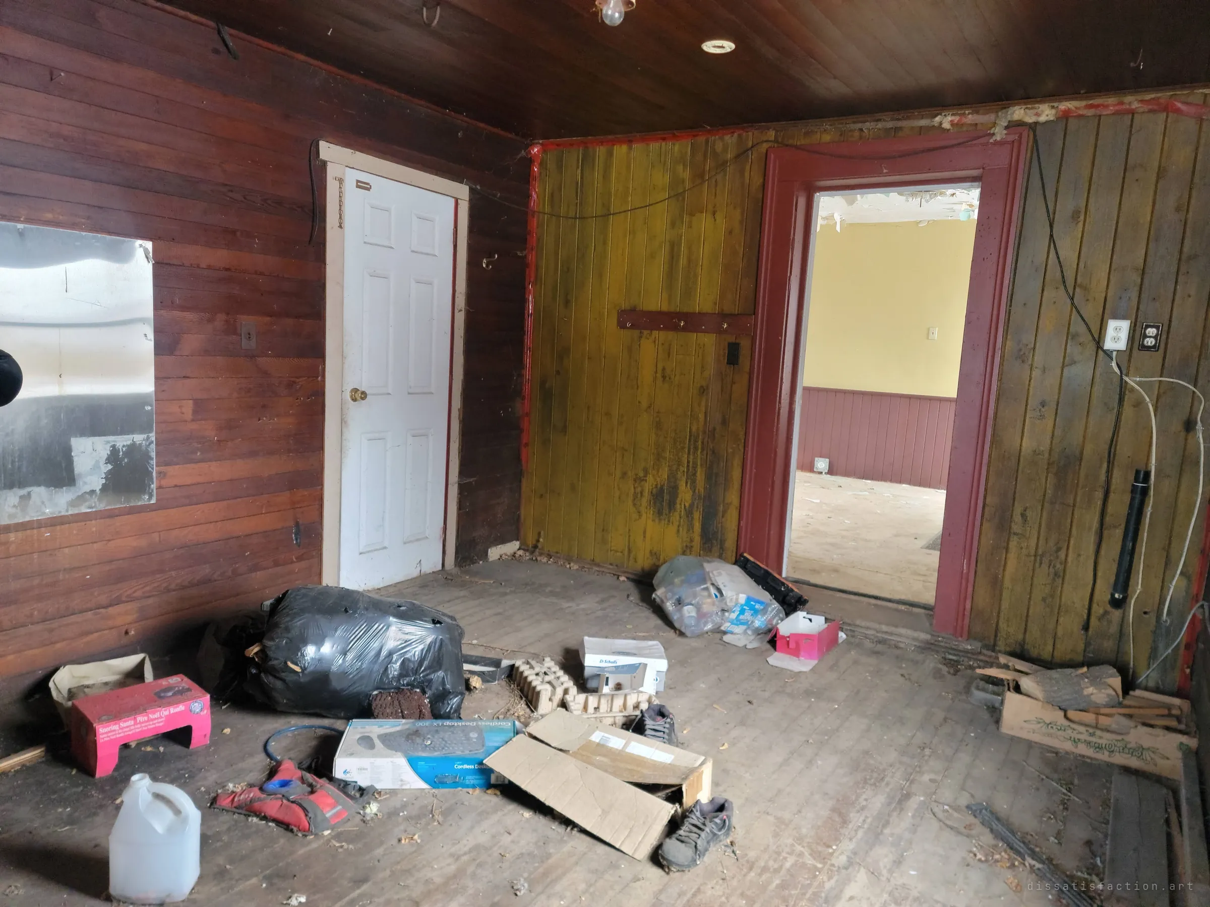 A wood-panelled back room littered with bags, boxes, old shoes and a jug, a mirror still hanging on the wall. Through the red doorway the yellow front room shows.