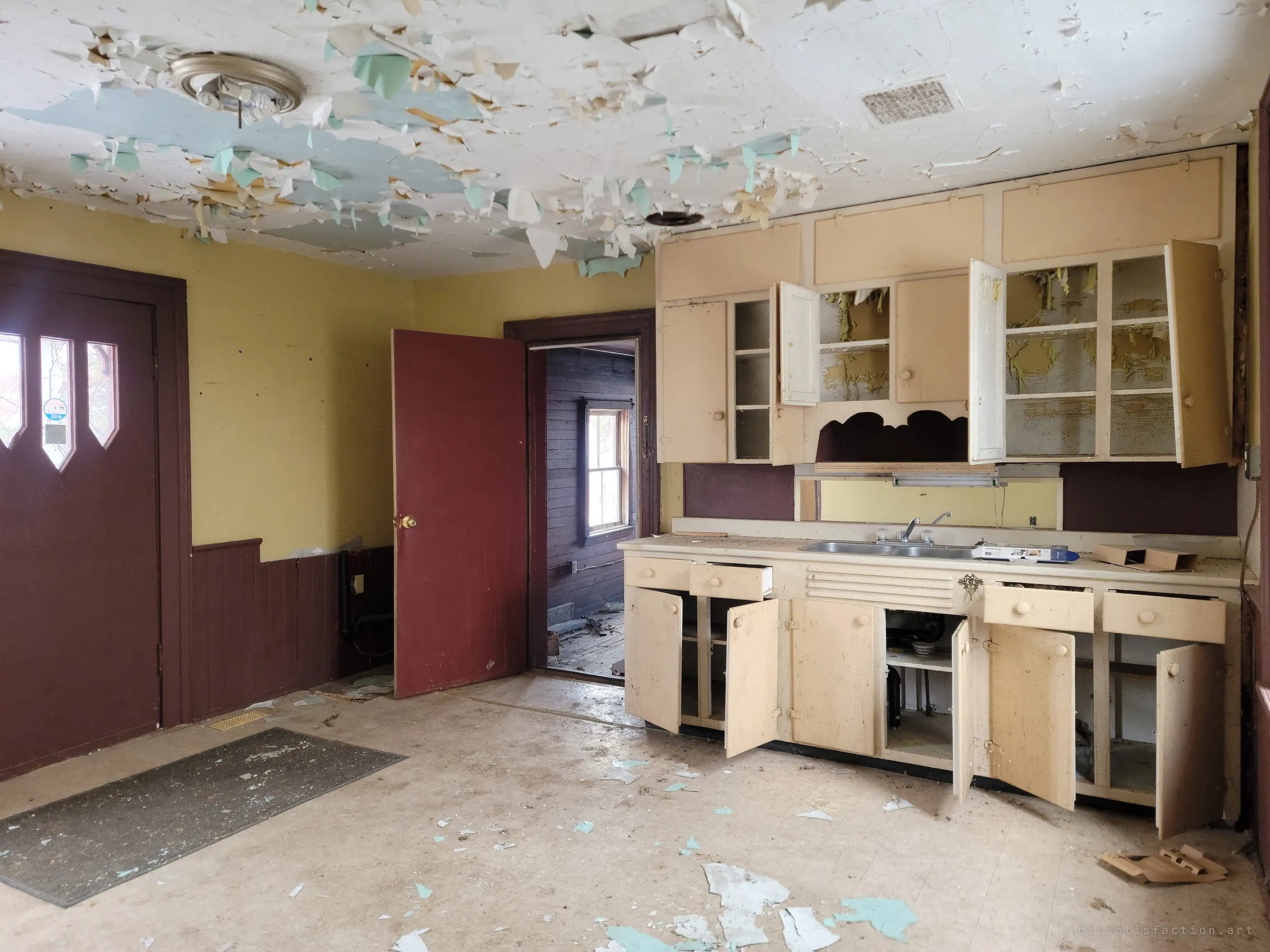 The kitchen: cream cabinets flung open and emptied, the ceiling peeling in curls of white and teal. A side door leads into a dark wood-lined back room.