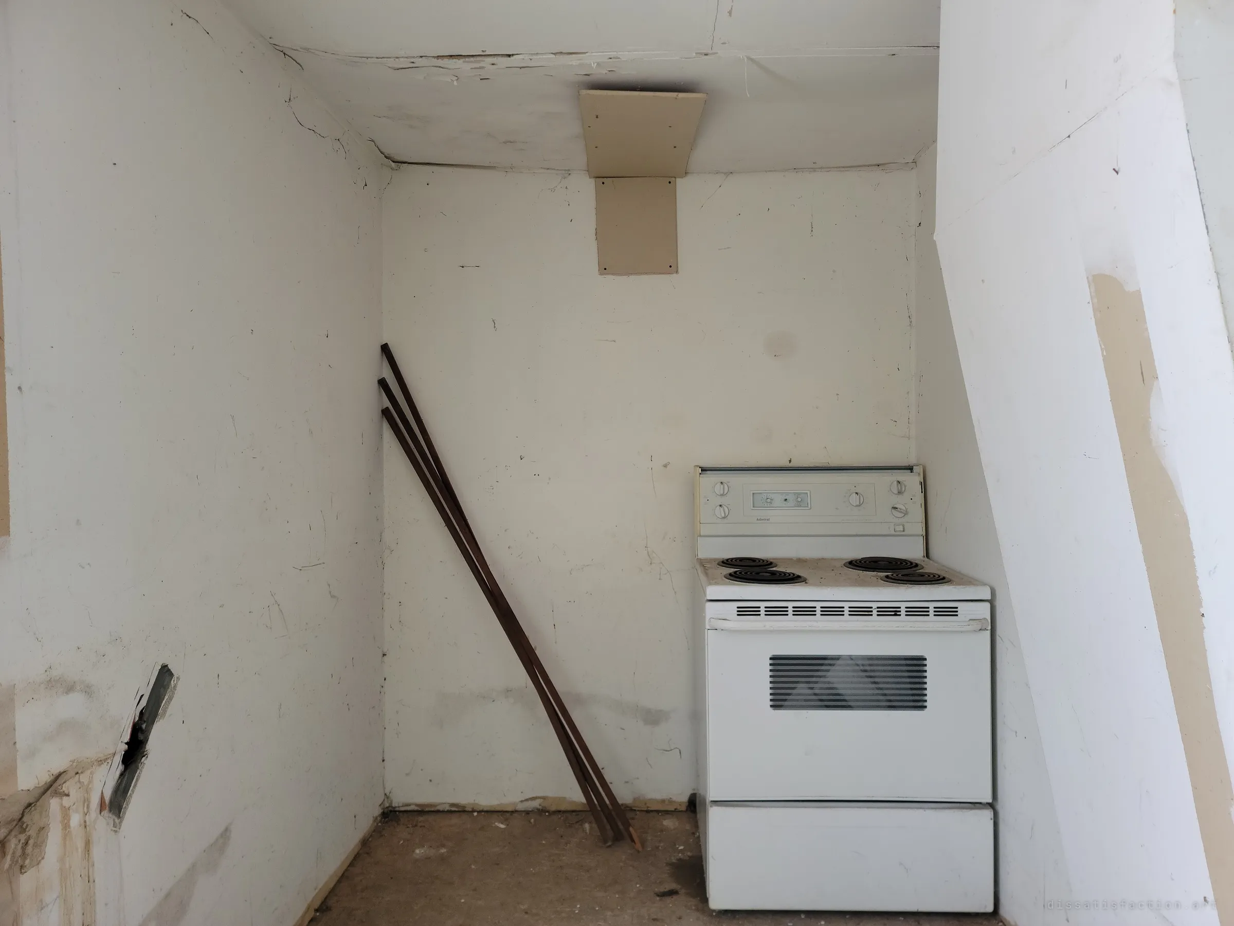 A white electric range left in a bare nook under the stairs, a few wooden slats leaning against the wall beside it.