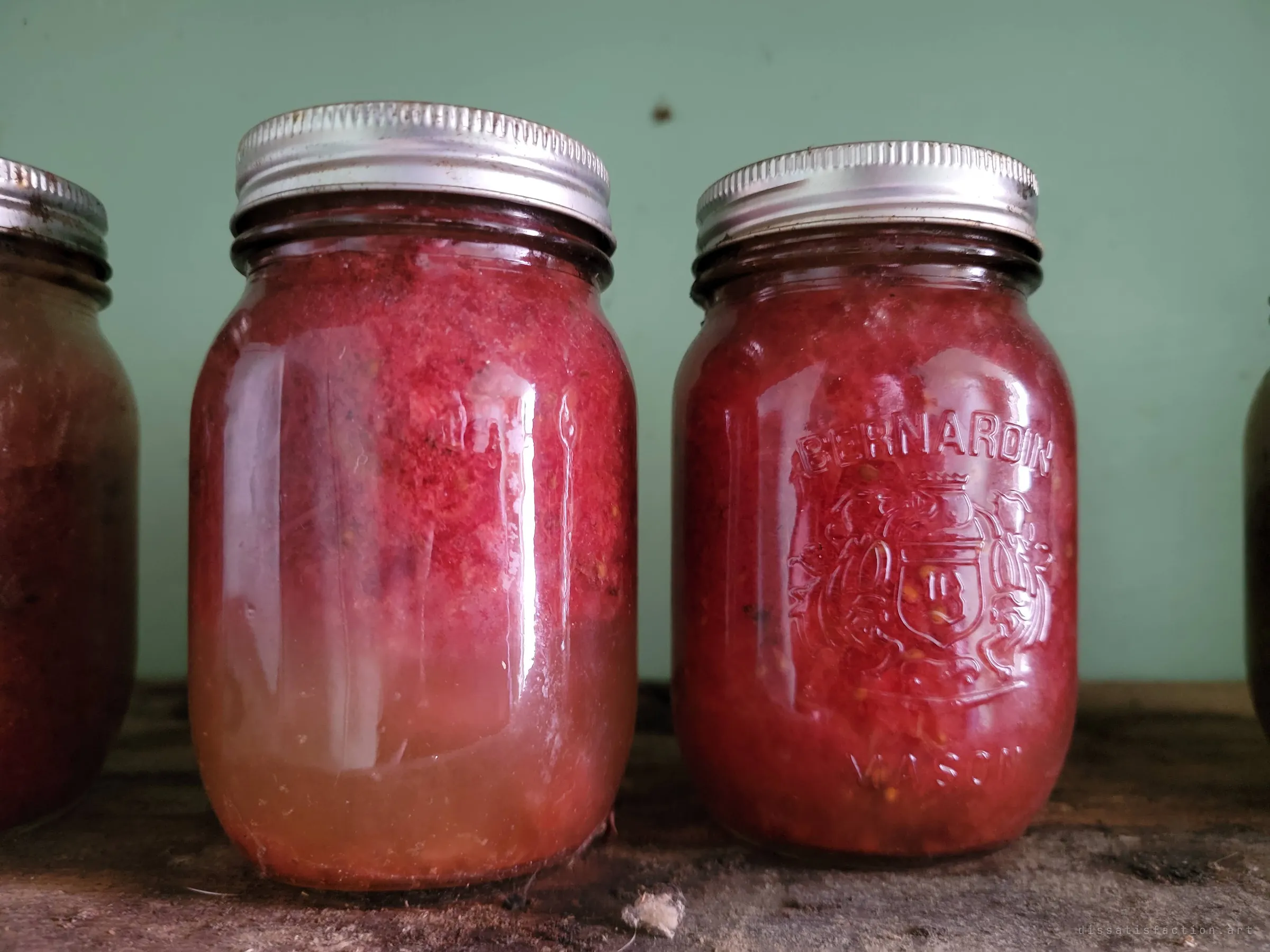 Close on two of the preserve jars, the red fruit separated into pulp and pale syrup behind embossed glass.