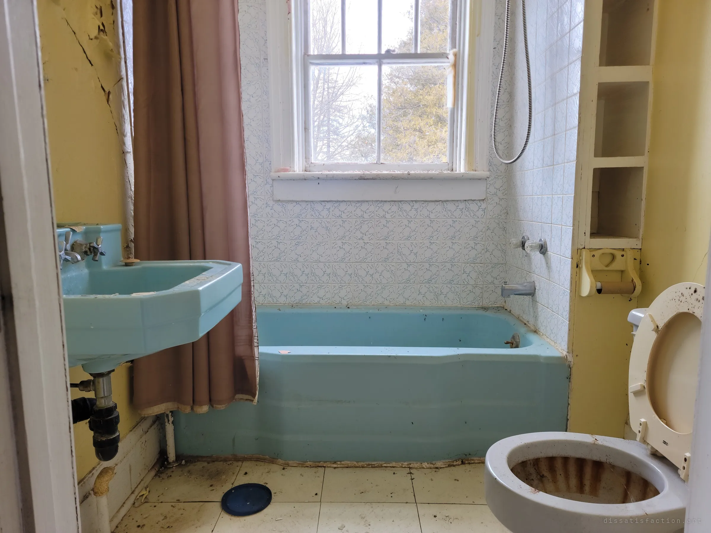 A small bathroom with a turquoise sink and tub, patterned tile and a window onto bare trees and a cedar. The toilet bowl is ringed with rust.