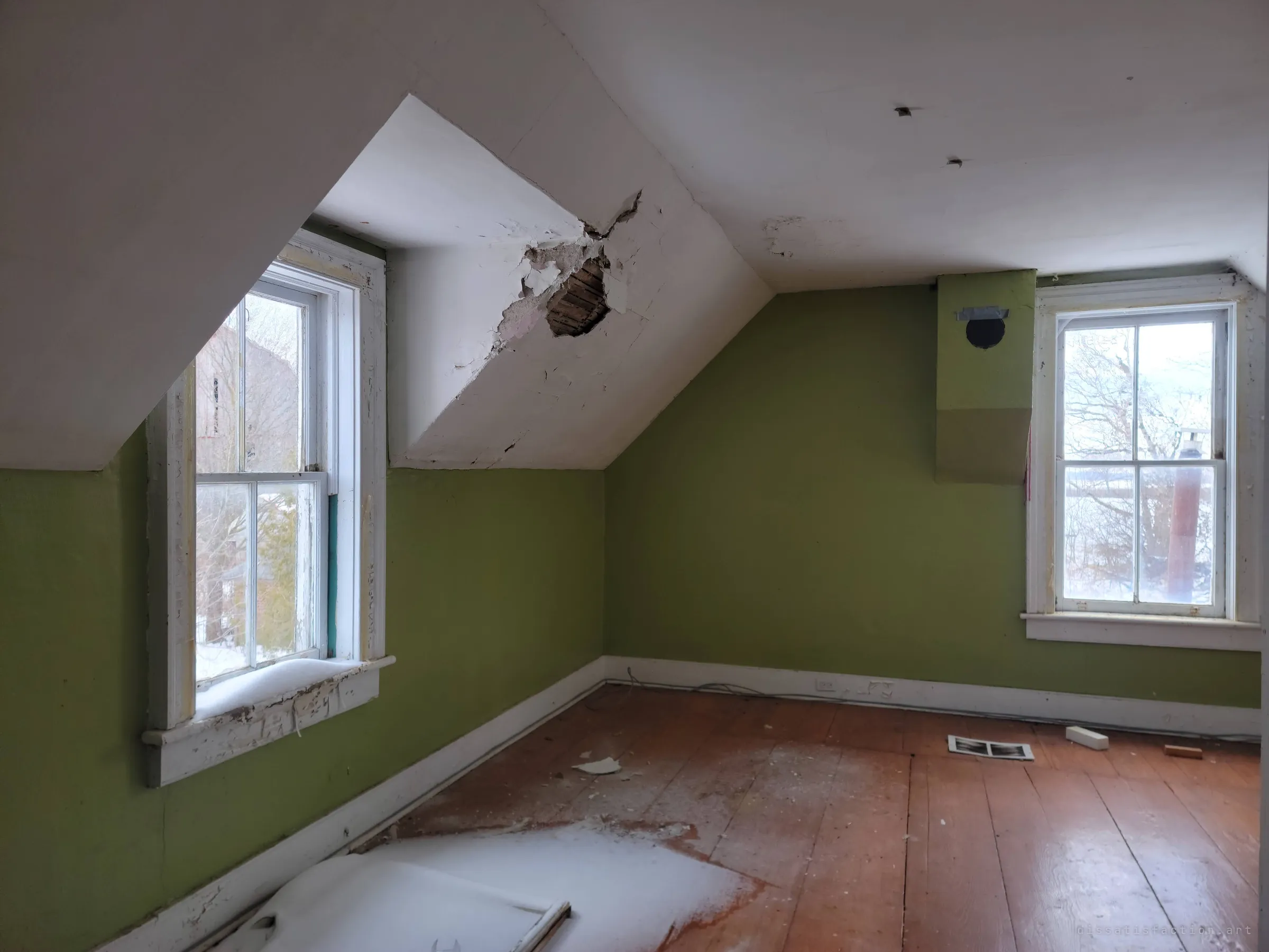 An attic bedroom with lime-green walls and a sloped white ceiling cracked open to the lath, snow drifted in across the plank floor. Winter light through two sash windows shows how exposed the upper floor has become.