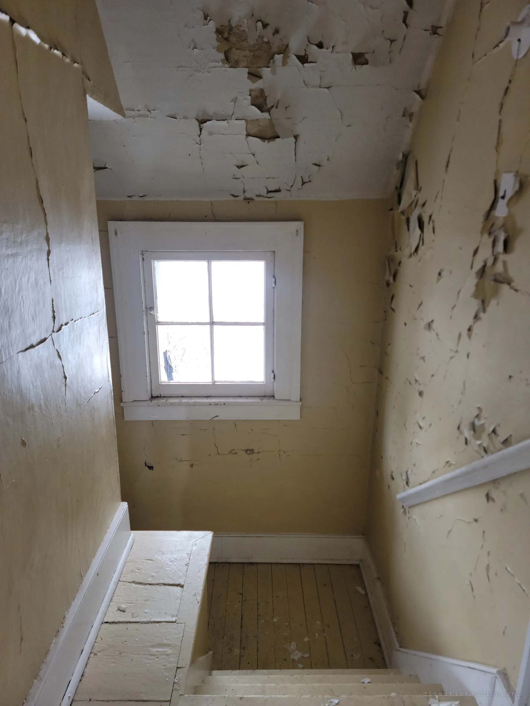 Looking down a narrow stairwell where yellow paint peels off the walls and ceiling in curled sheets toward a small four-pane window. The decay of the house reads in every surface.