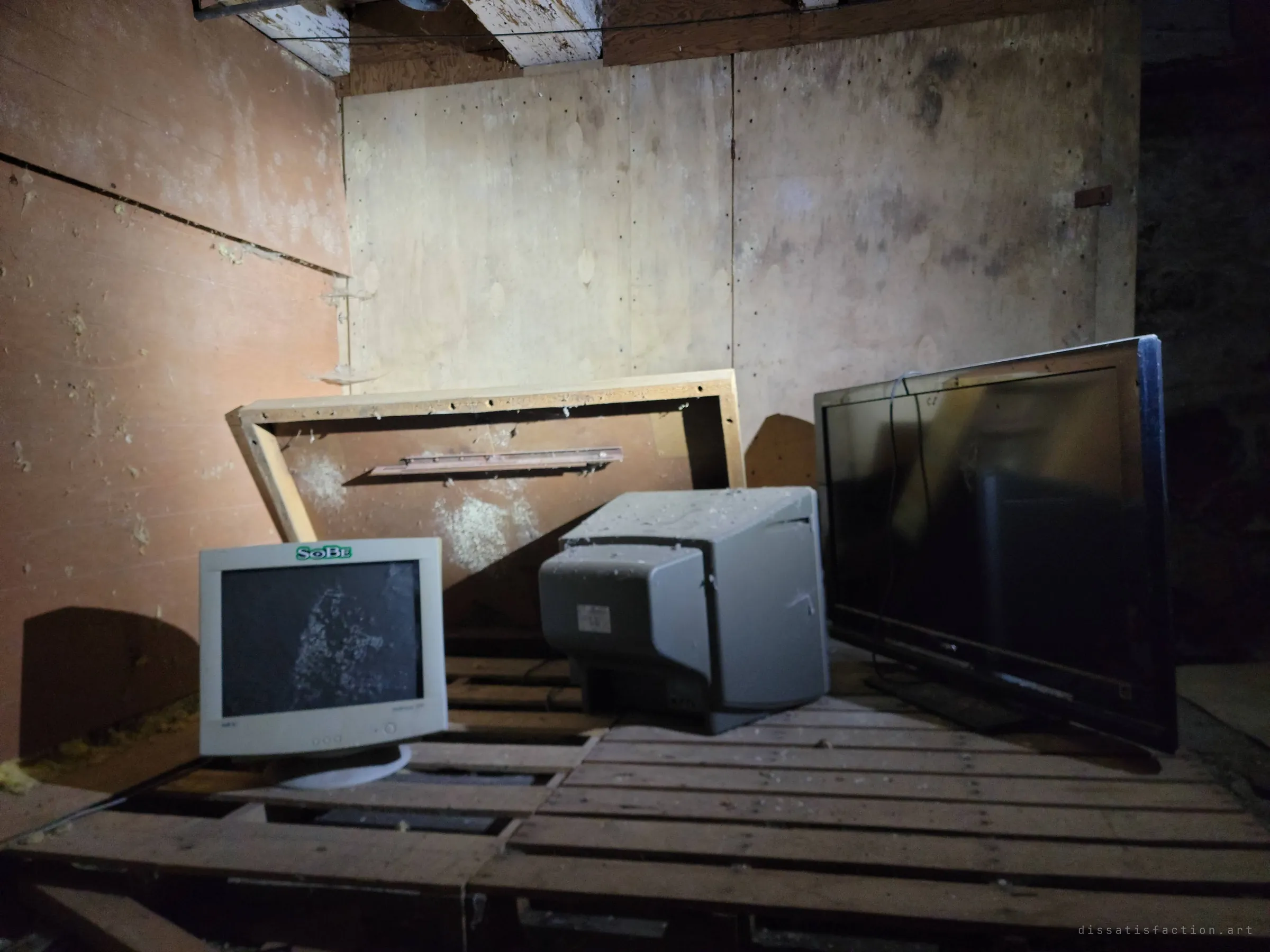 A flashlit corner of a plywood-lined loft holding an old beige CRT monitor with a cracked screen, a second monitor turned away and a flat-screen TV, all on pallets under dust and insulation. Obsolete electronics left behind like a small shrine.