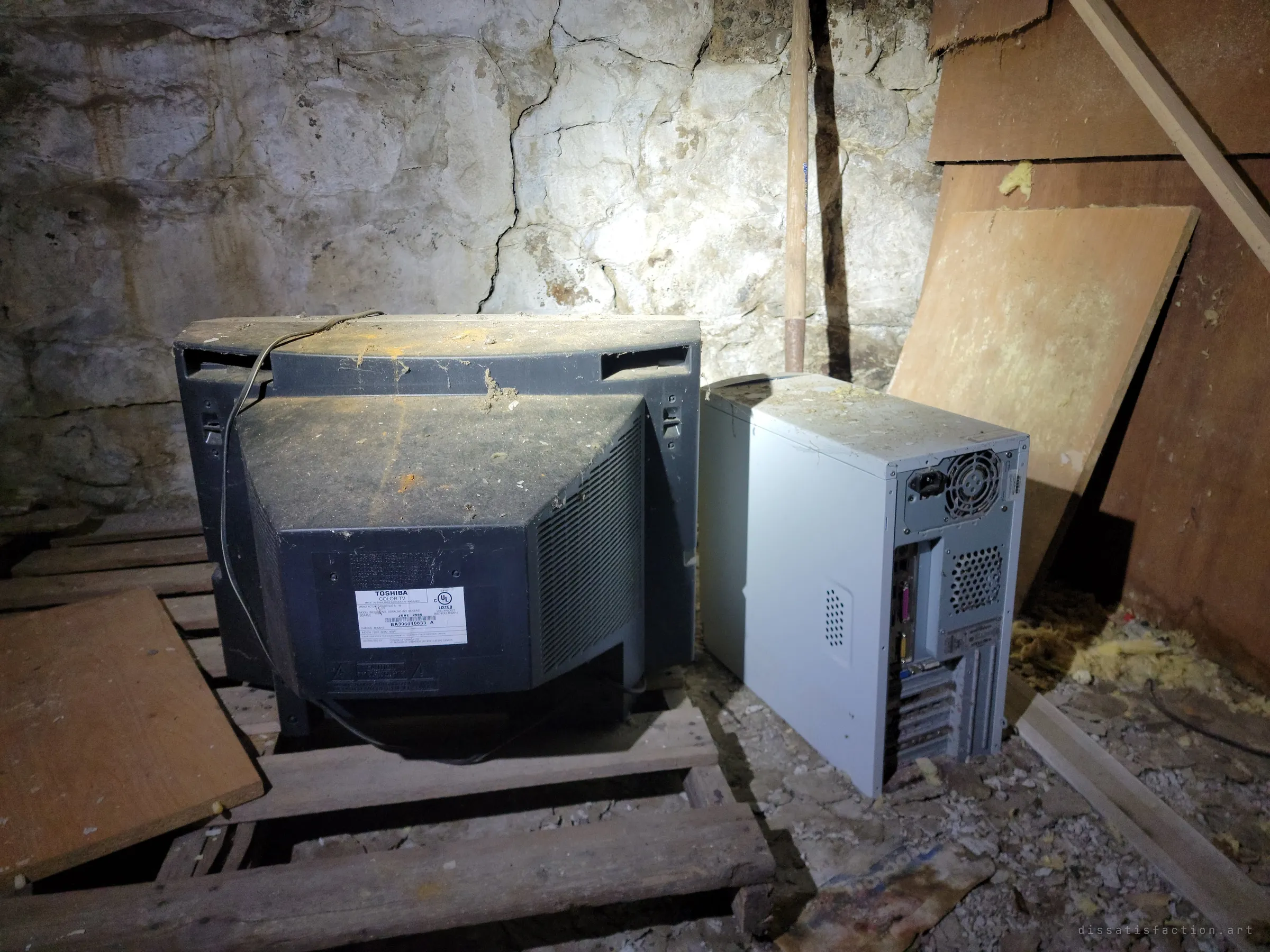 The back of a bulky CRT television and a grey desktop tower sit on pallets against a rough stone foundation wall, lit by a flashlight. Early-2000s tech abandoned in a cellar.