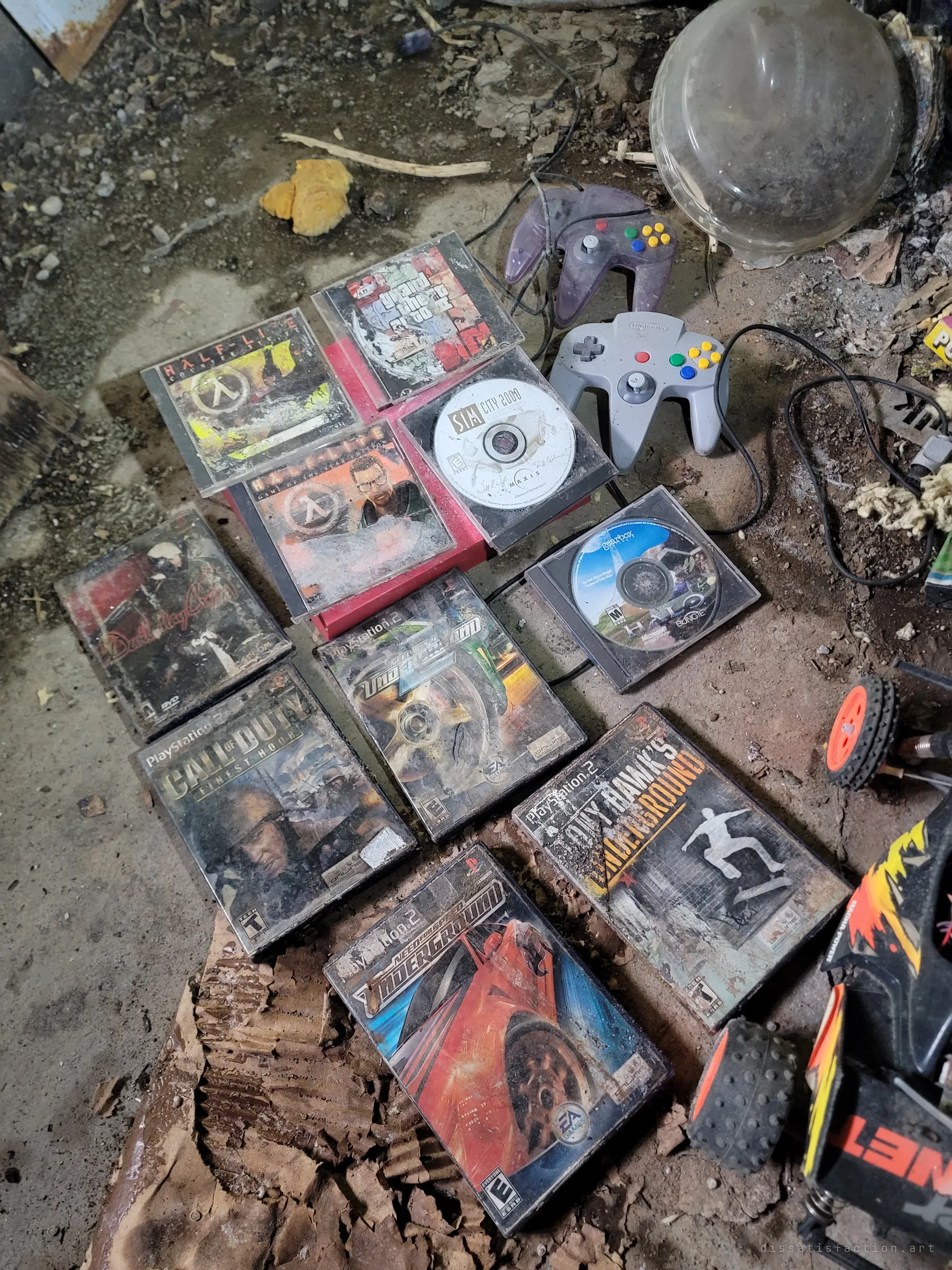 A spread of grimy PC and PlayStation 2 game cases, two N64 controllers and a toy RC car lie on a dirty floor. A childhood game collection left to rot.