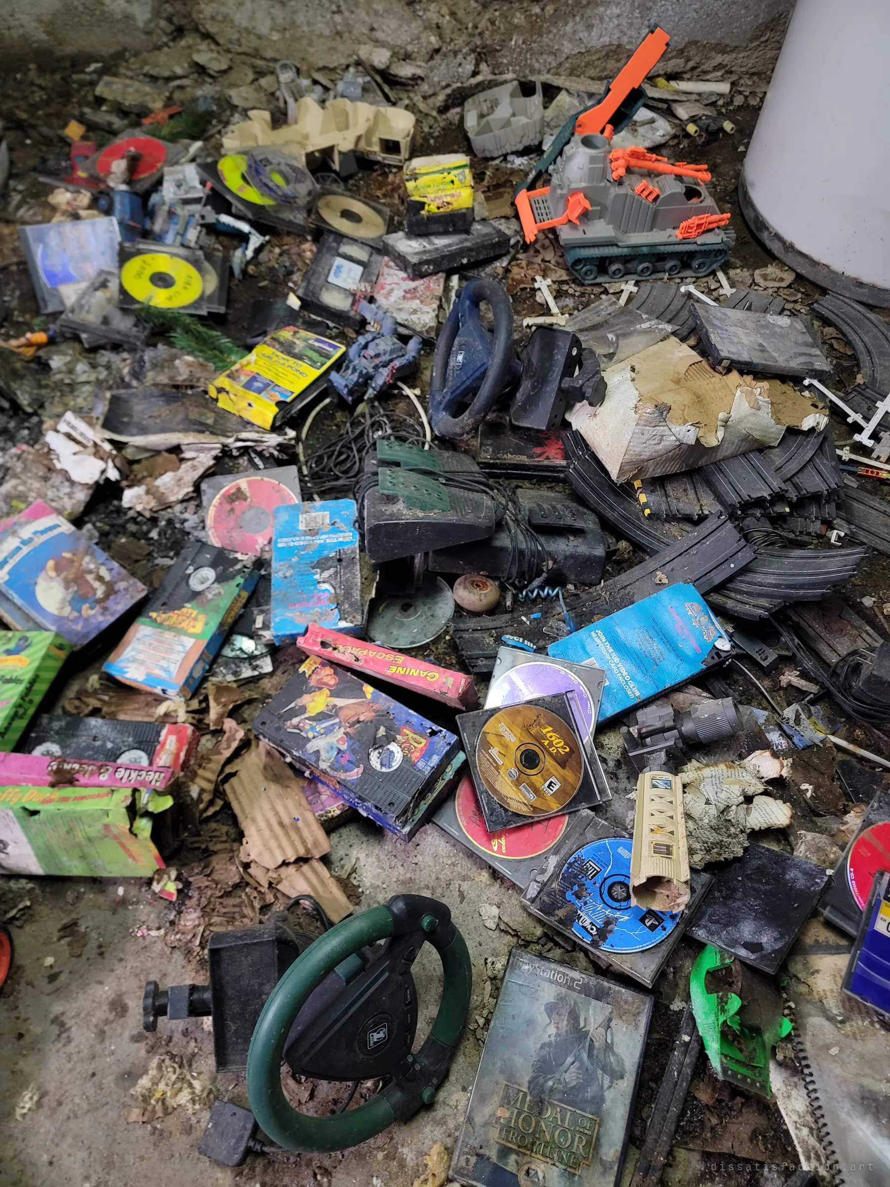 A floor heaped with kids' VHS tapes, game discs, racing-wheel controllers, slot-car track and a toy tank, all soaked and filthy. The pile captures a whole family's era of entertainment in one heap.
