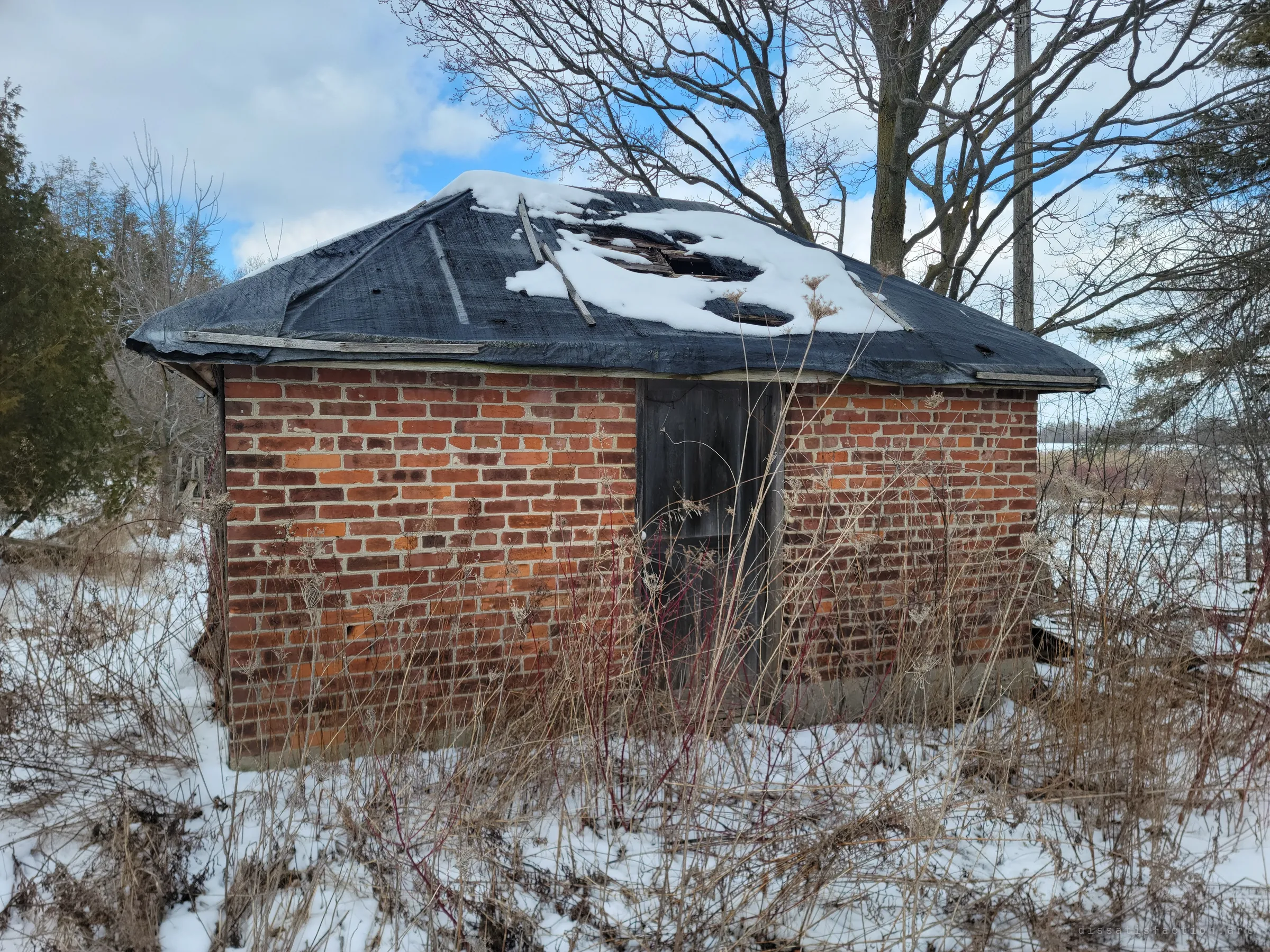 A small brick outbuilding with a tarp-covered hip roof, its snow-laden top caved in, stands among dry weeds and snow. The weather is slowly getting inside.