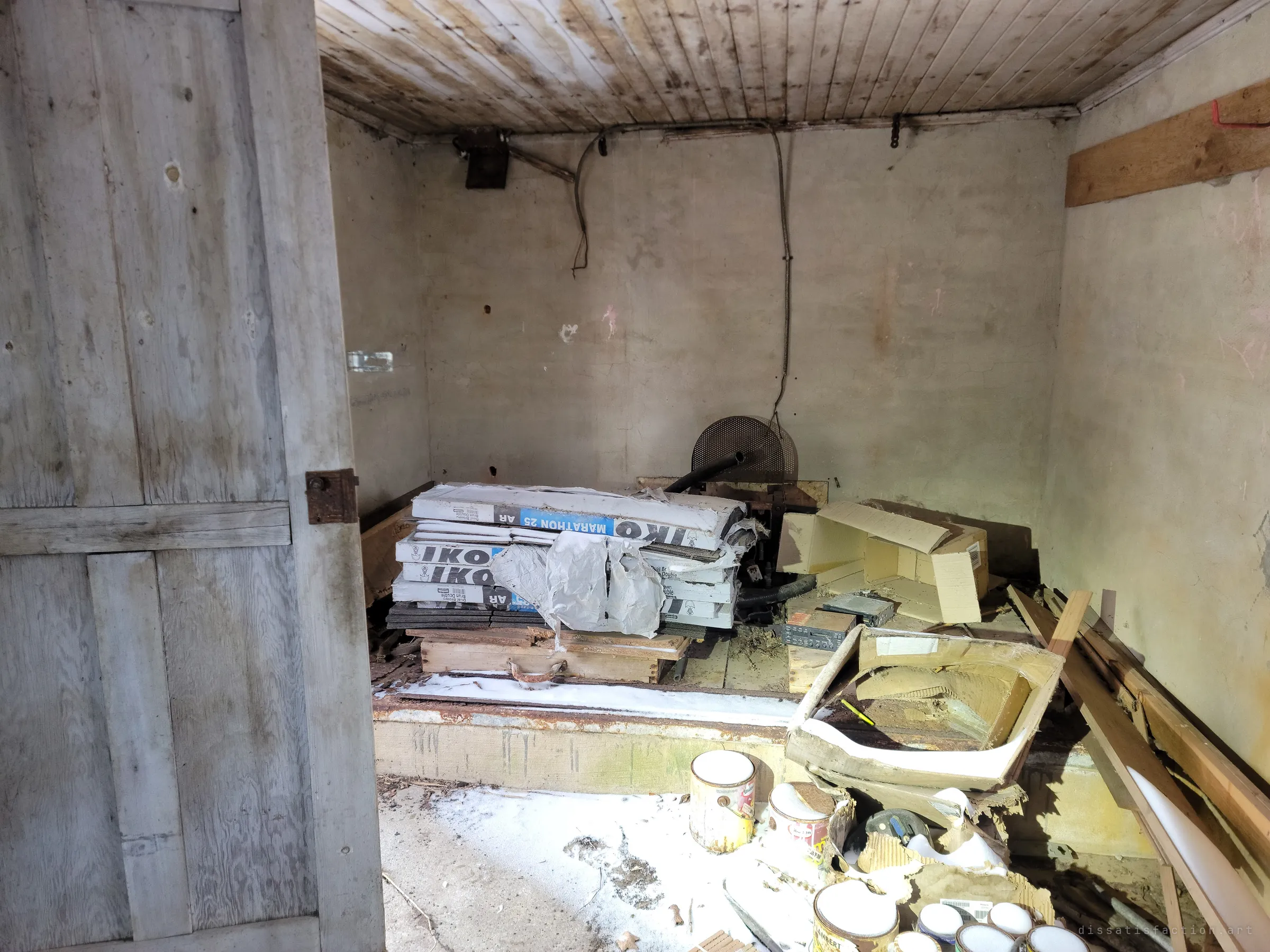 Inside the outbuilding, bundles of roofing shingles, old paint cans and a broken sink sit on a raised concrete ledge beneath a stained board ceiling, snow dusted across the floor. Leftover renovation supplies that never got used.