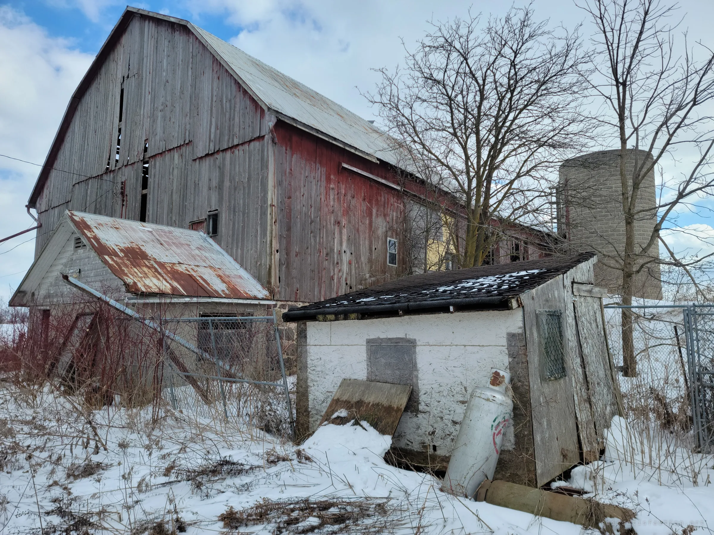 The big red-and-grey barn rises behind a small whitewashed pump house with a propane tank leaning on it, a concrete silo standing beside bare trees. The farmyard's working heart, now silent.