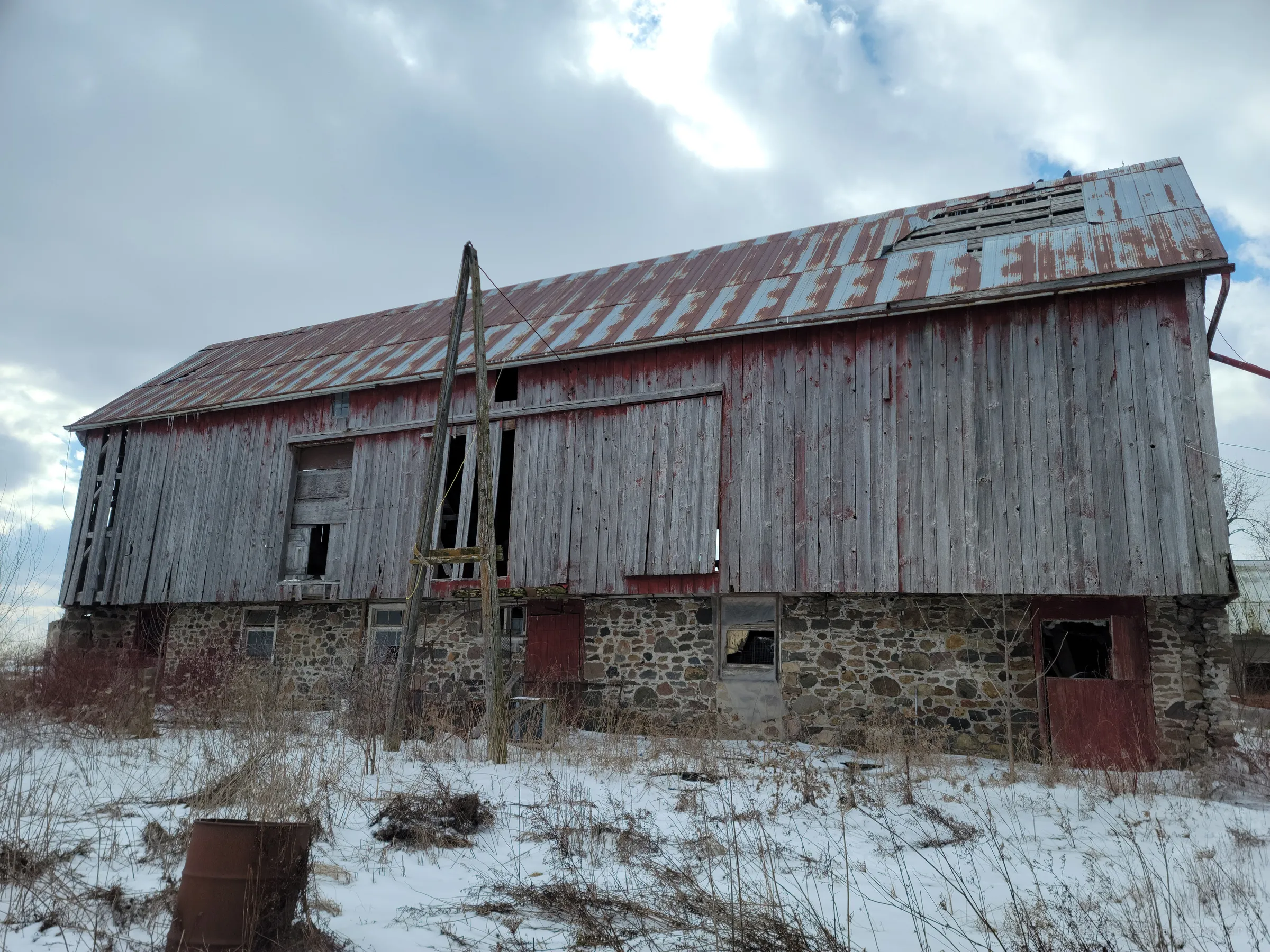 A long bank barn with weathered grey board siding, a rust-streaked tin roof missing panels, and a fieldstone foundation half buried in snow. The leaning utility pole and gaps in the boards show how far the structure has gone.