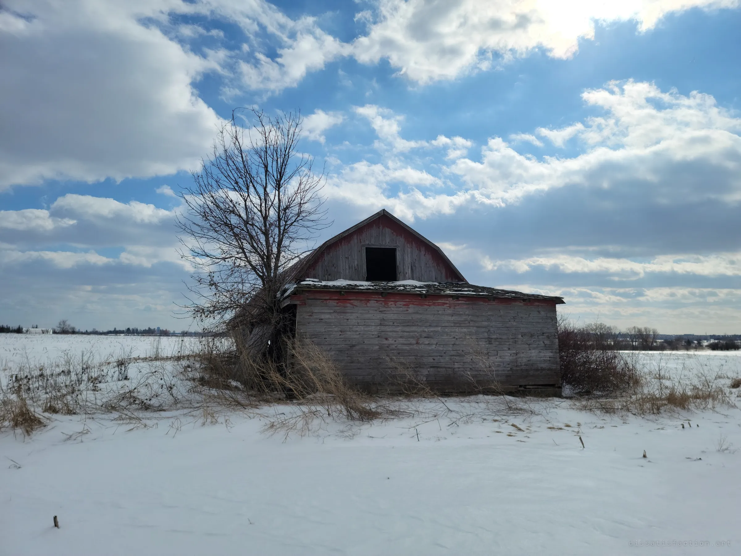 A small red-trimmed gambrel outbuilding sits alone in a snow-covered field beside a bare tree under a bright, broken sky. It shows the farm's scattered outbuildings left to the weather.