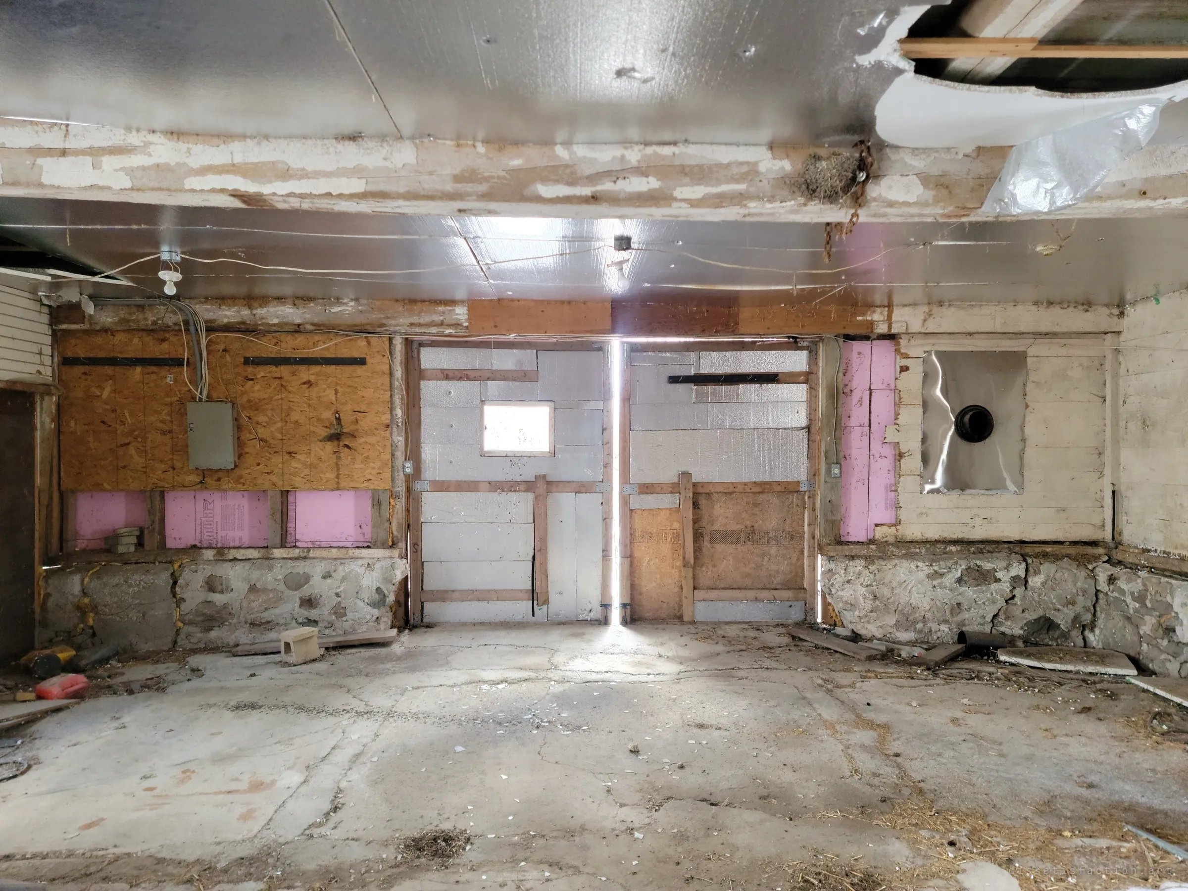 A gutted garage bay with foil-faced insulation, patched plywood and pink foam board, and a thin line of daylight splitting the closed double doors. Loose wiring and a bird's nest hang from the beams.