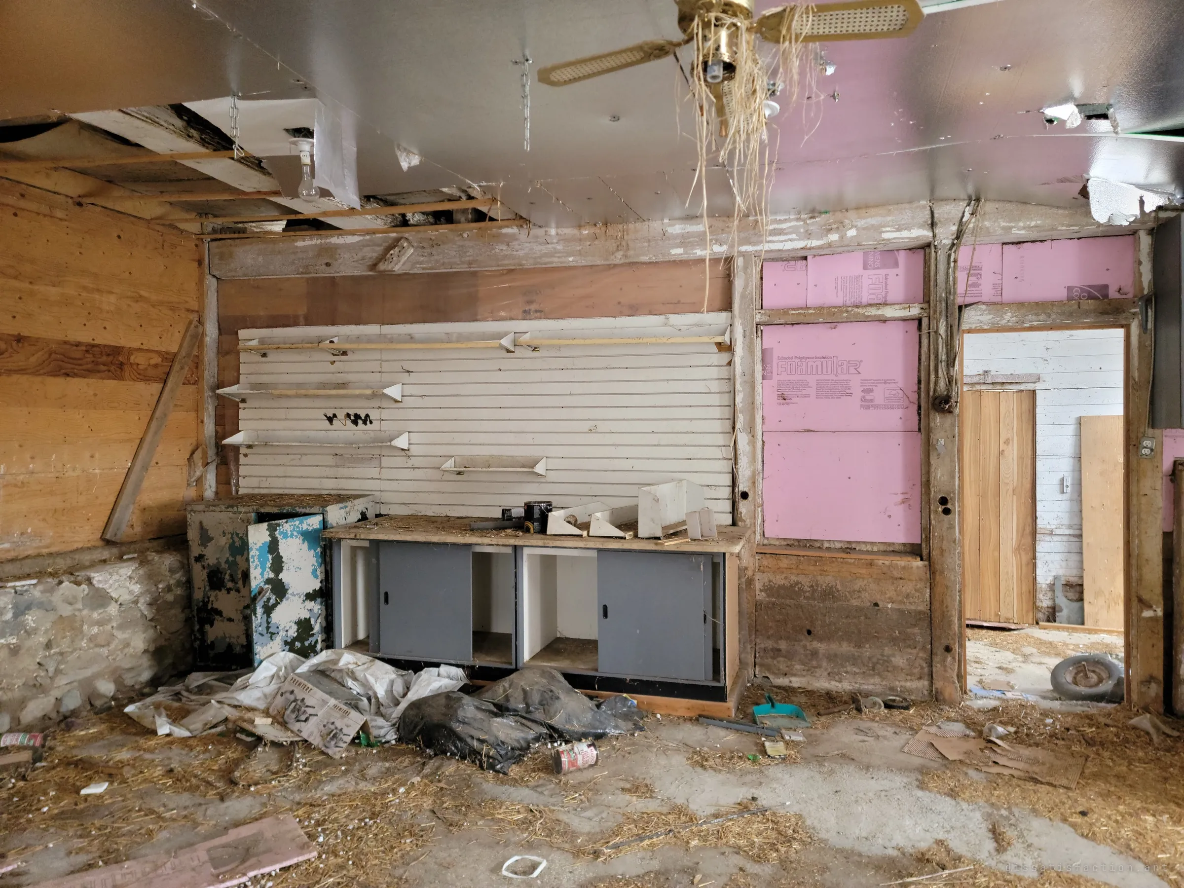 A workshop corner with slatwall shelving, a peeling blue metal cabinet and grey sliding-door benches above straw and tarps on the floor. A brass ceiling fan hangs tangled in straw, and a doorway leads to another whitewashed room.