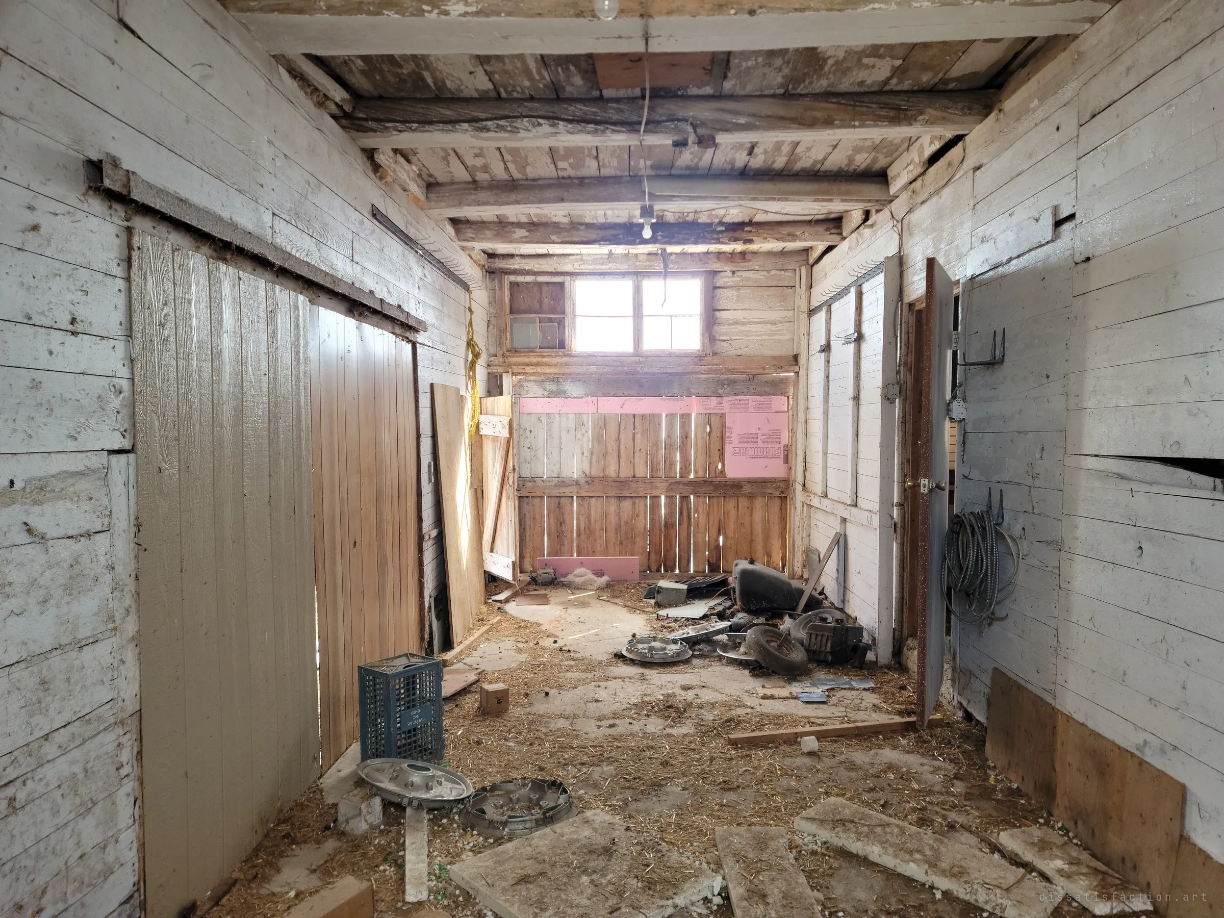 A narrow whitewashed room with sliding board doors on one side and a window at the far end lighting a slatted wall and piles of hubcaps and parts. Straw and broken foam cover the floor.