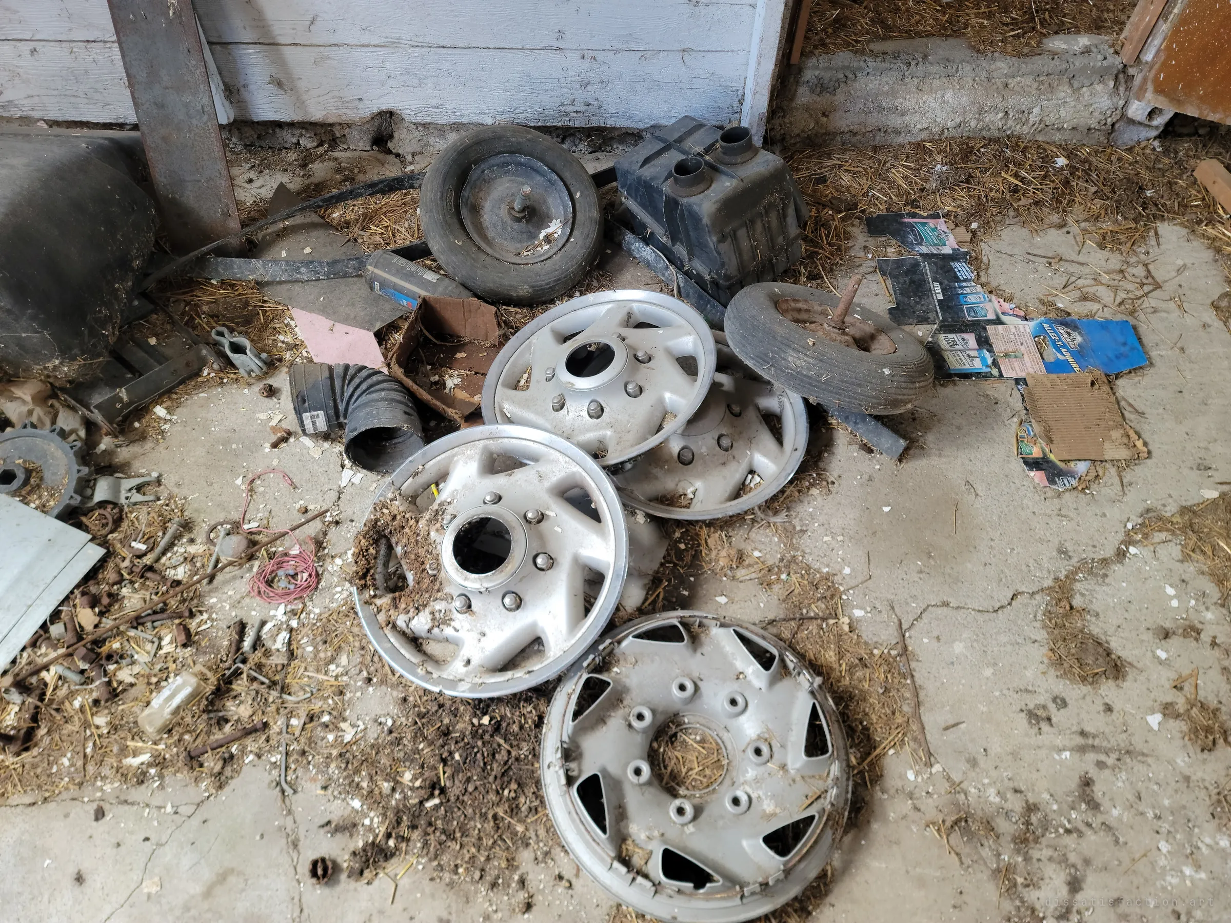 A pile of dusty silver hubcaps, small wheelbarrow tyres and a plastic tank on a cracked concrete floor strewn with straw and torn packaging. A close look at what was left behind in the barn.