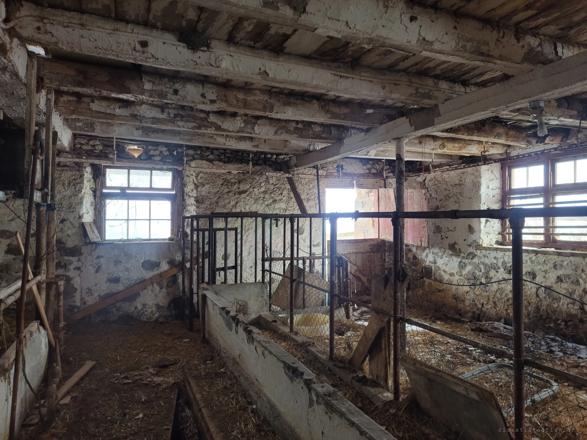 A stone-walled stable room with multi-pane windows, rusted pipe stalls and chain-link pens lit by pale winter light. The whitewash is peeling off both the walls and the low timber ceiling.