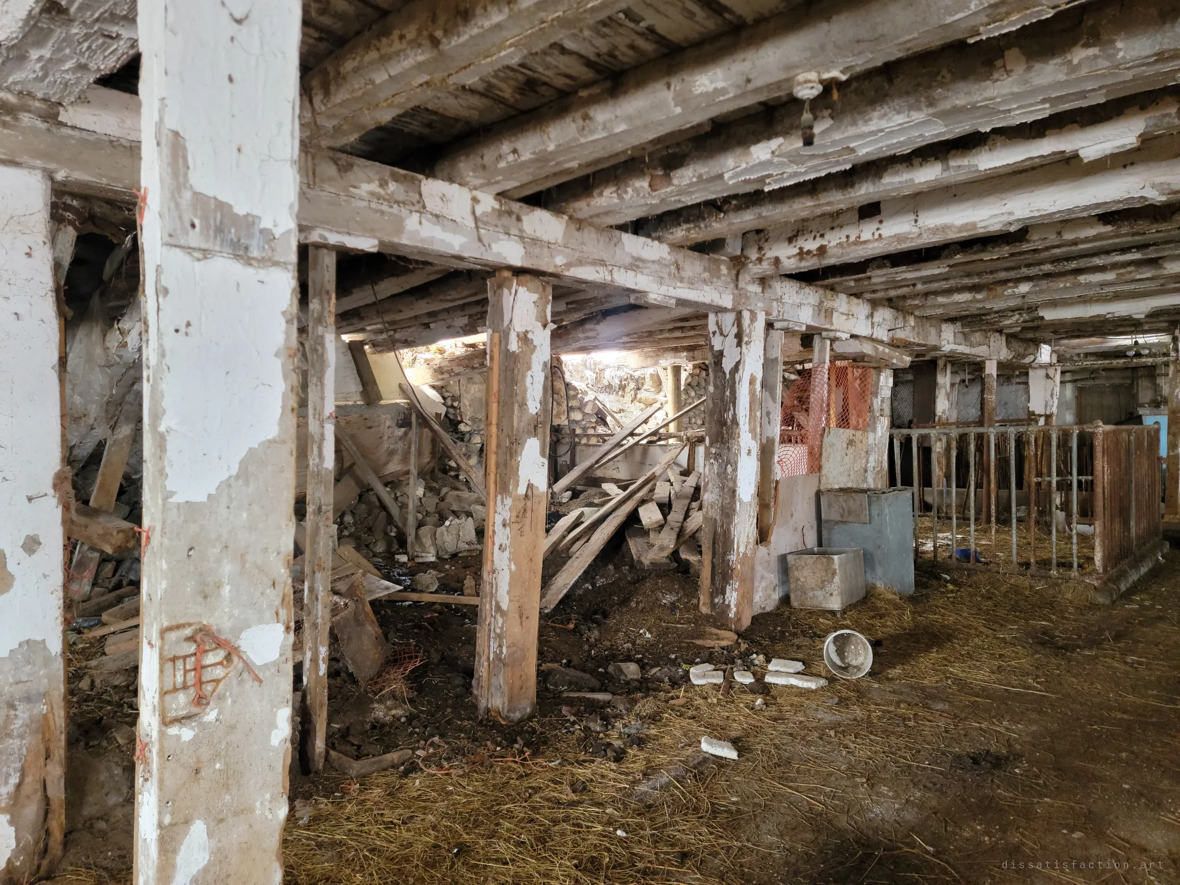 Whitewashed timber posts hold up a sagging floor, while behind them part of the stone foundation has collapsed into a heap of rubble and broken boards. Orange safety mesh and a rusted pen sit off to the side.