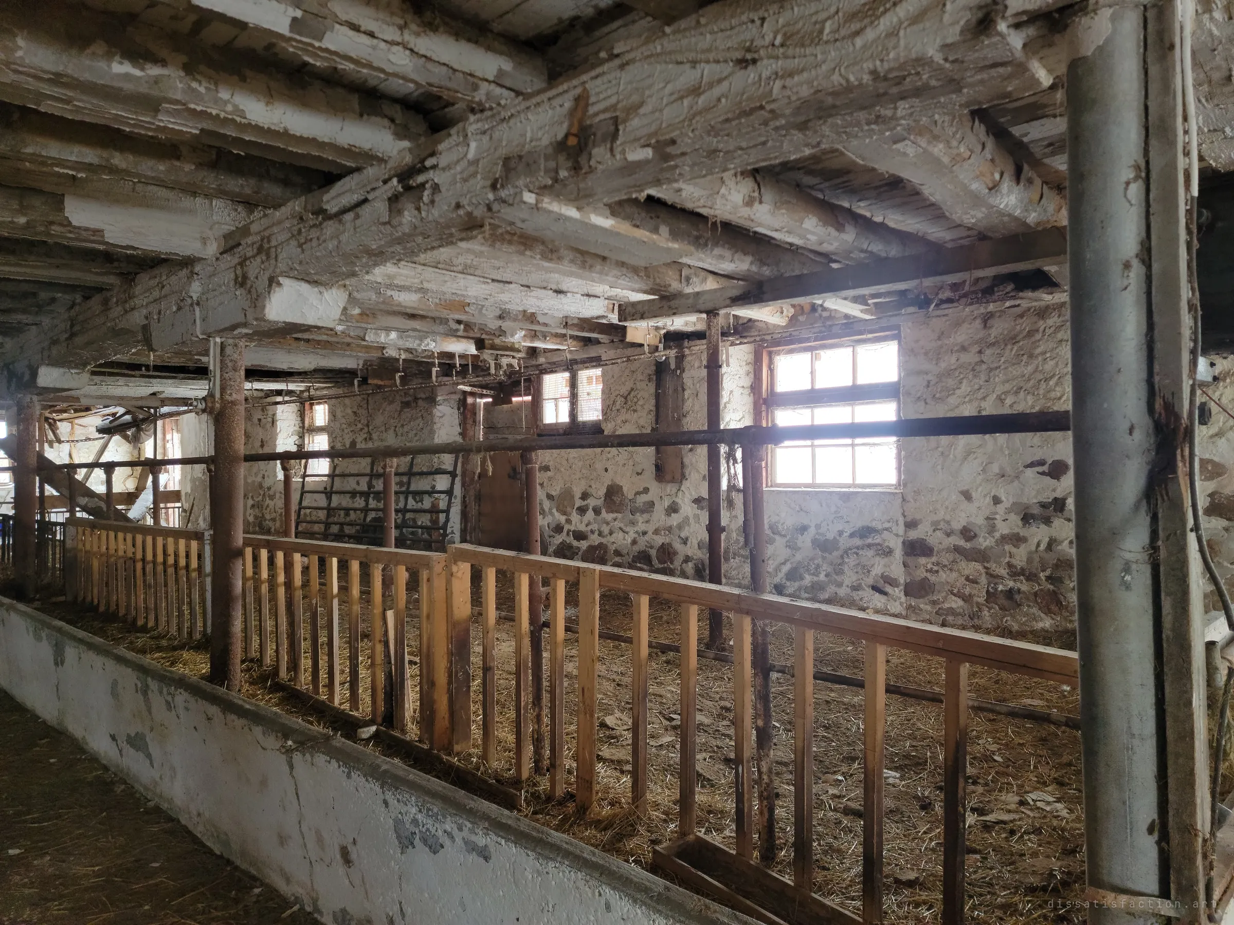 Rough-hewn beams with flaking white paint span a stable lined with new wooden rails and a long concrete trough. Light from the windows in the fieldstone wall falls across the straw.