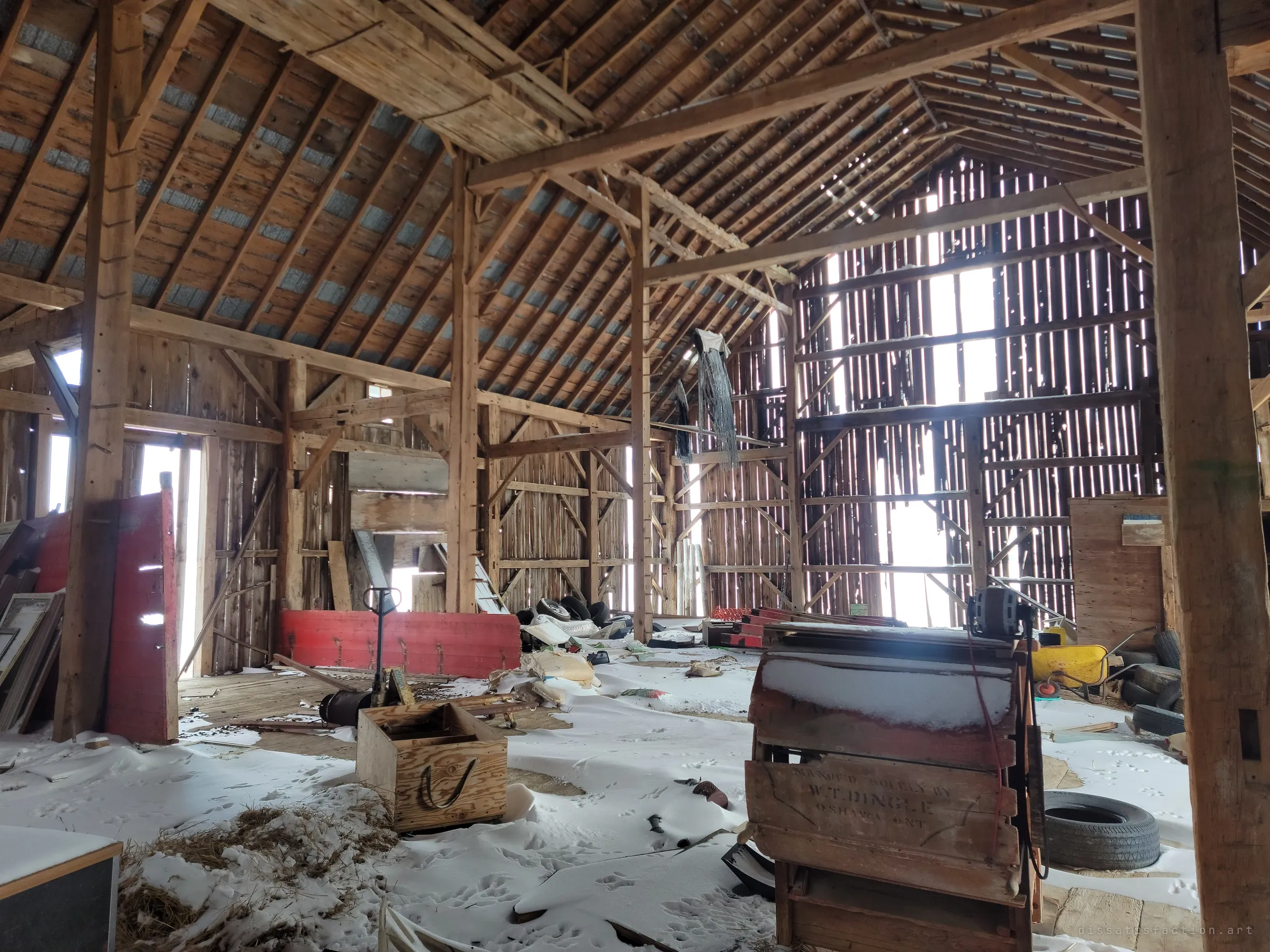 The main floor of a timber-frame bank barn under drifted snow, light slicing through the gaps in the far wall onto an old wooden fanning mill, crates and scattered tires. The hand-hewn frame and snow indoors show how open the building has become.