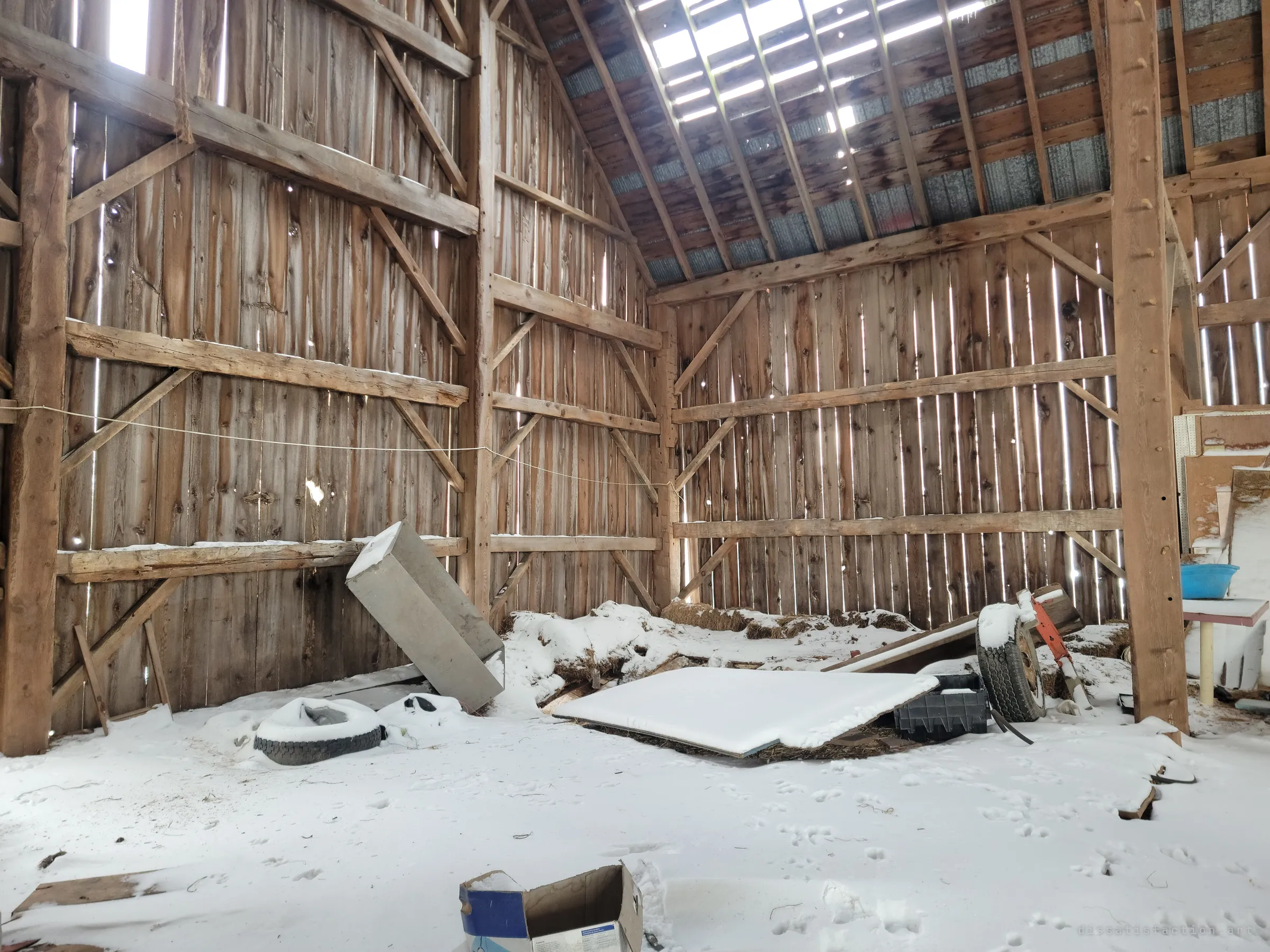 A snow-covered corner of the barn's hay mow, with light leaking through missing roof boards and the gaps in the plank walls. A tipped-over metal cabinet, a tire and a wheel lie half buried in the snow.