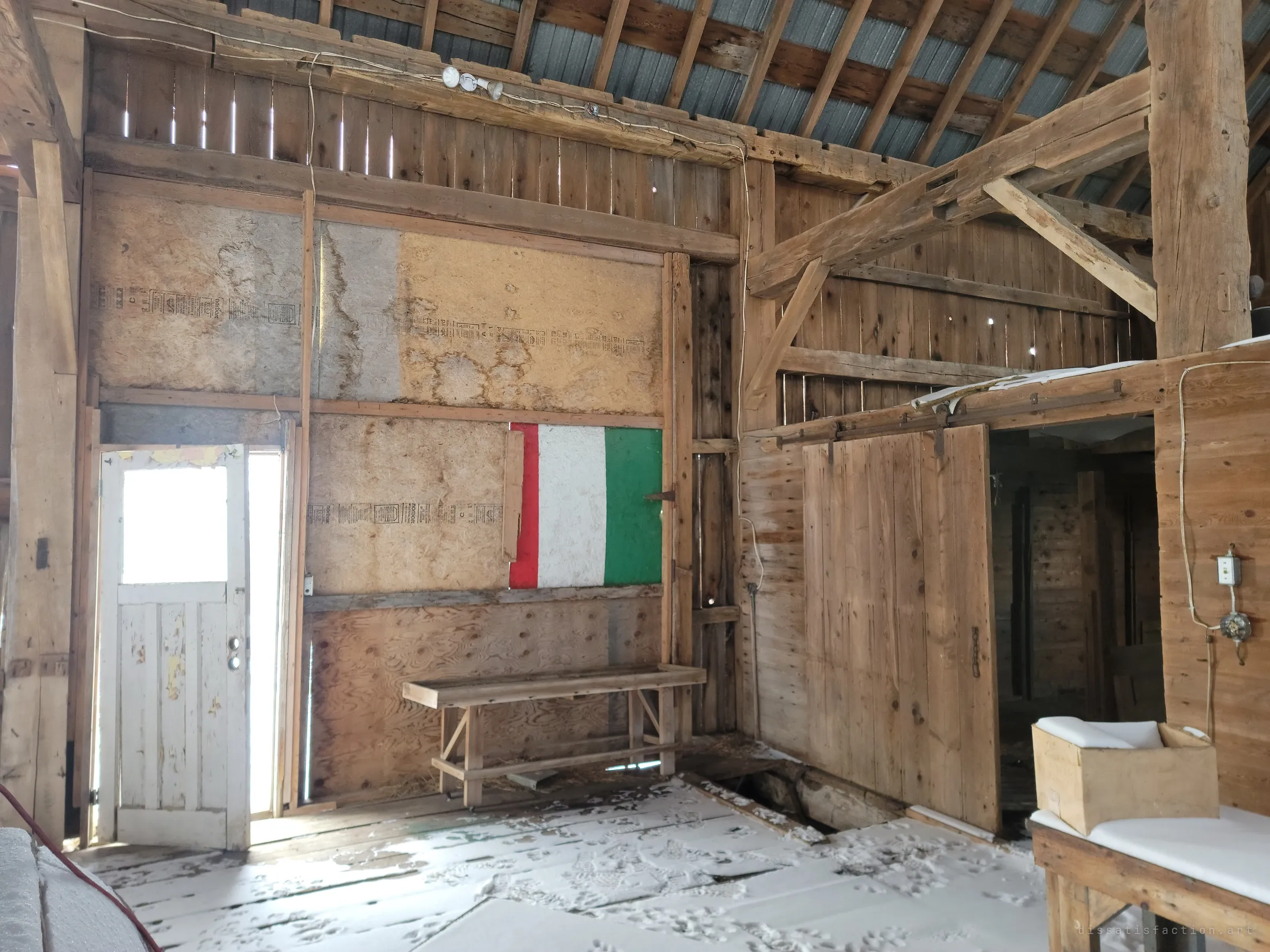 An insulated room built inside the barn, with a flaking white door, a red, white and green painted panel on the wall and a long wooden bench. Snow has blown across the floorboards and a sliding door opens into a dark stall area.