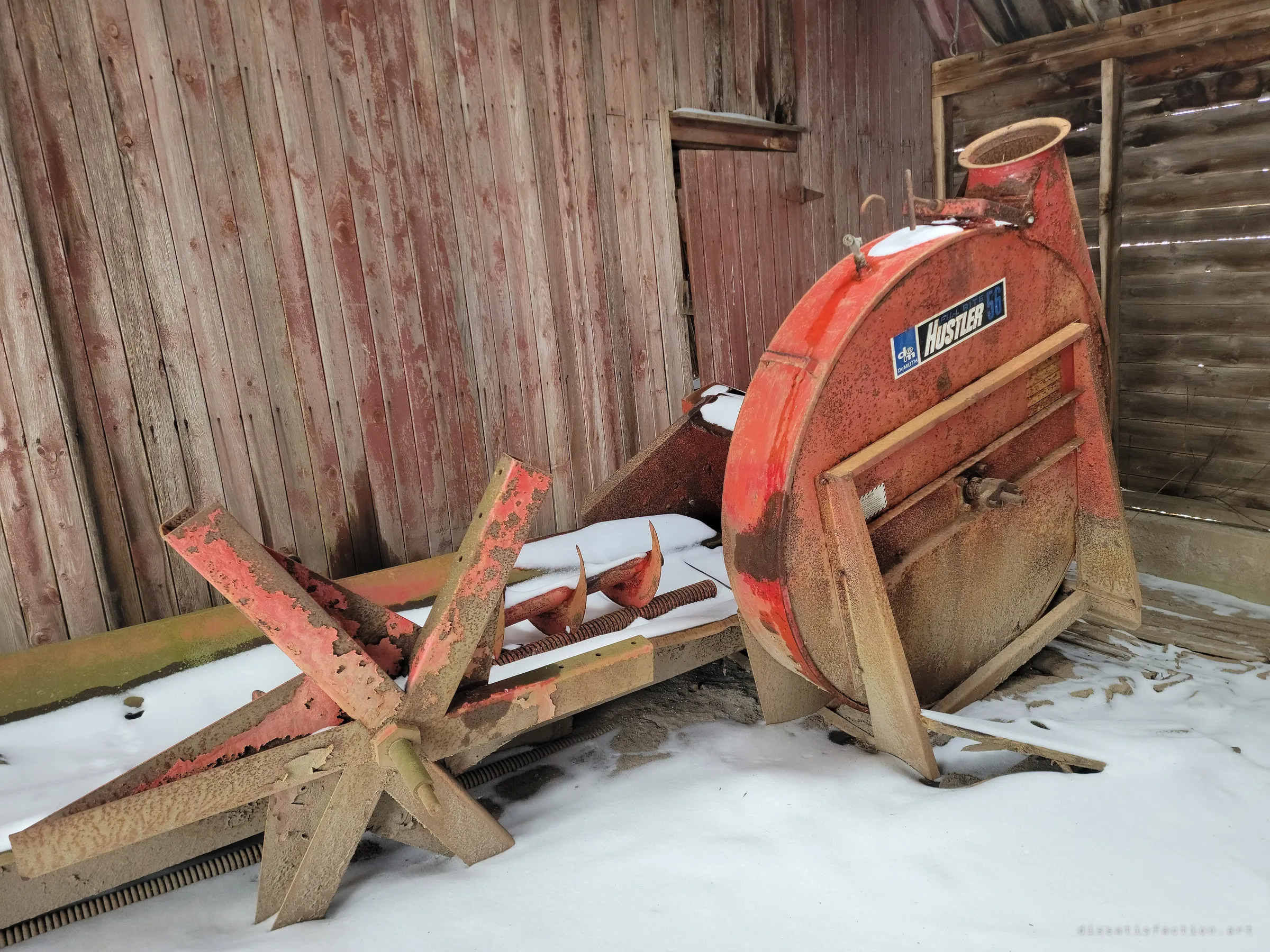 A rusting orange forage blower and its auger rest against a weathered red plank wall, dusted with snow. The farm machinery was left where it last worked.