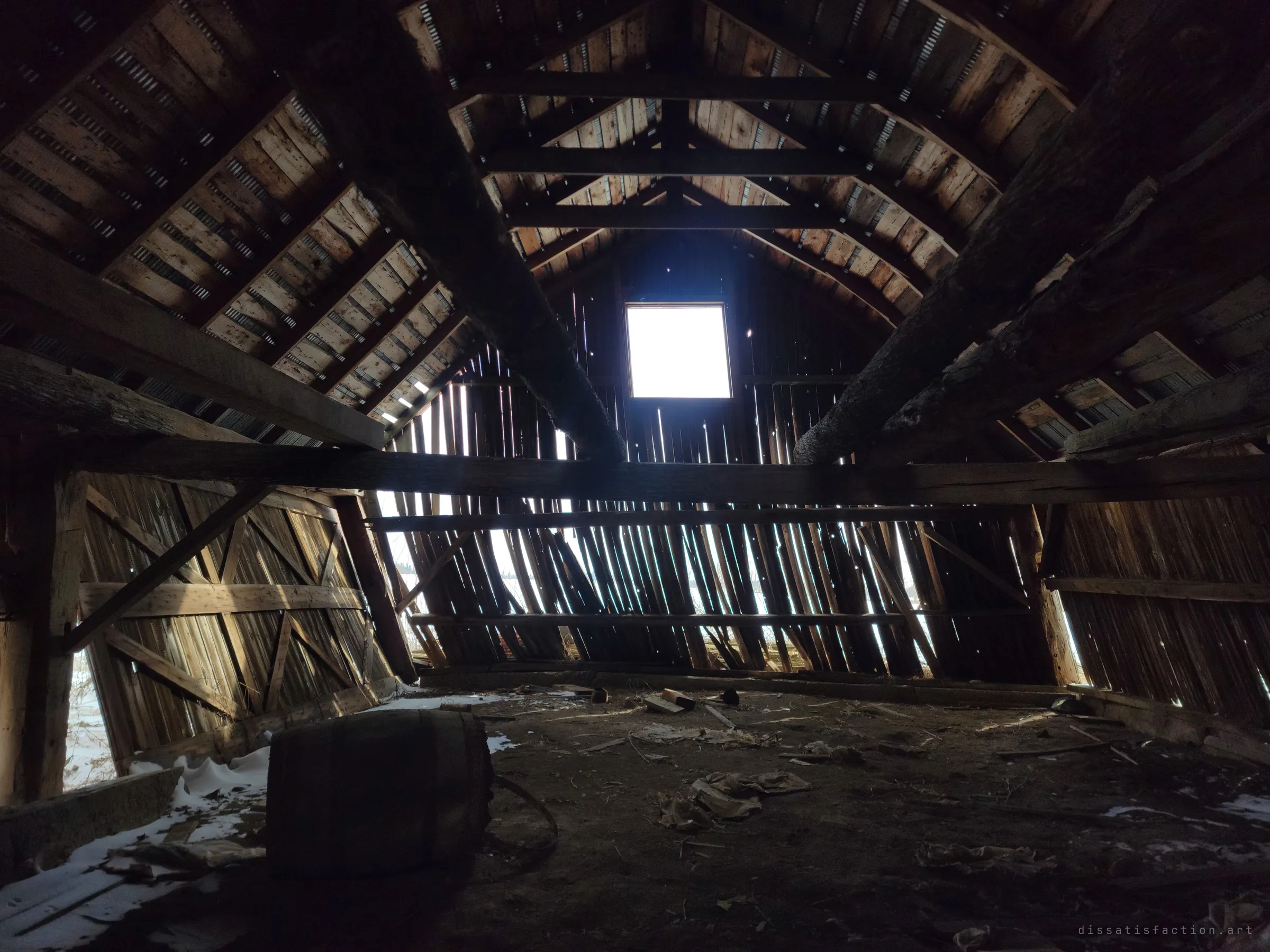 The dark upper loft of the barn, its curved gambrel rafters lit by a single square window at the gable end. An old wooden barrel lies on its side on the dirt-covered floor while the walls lean outward.