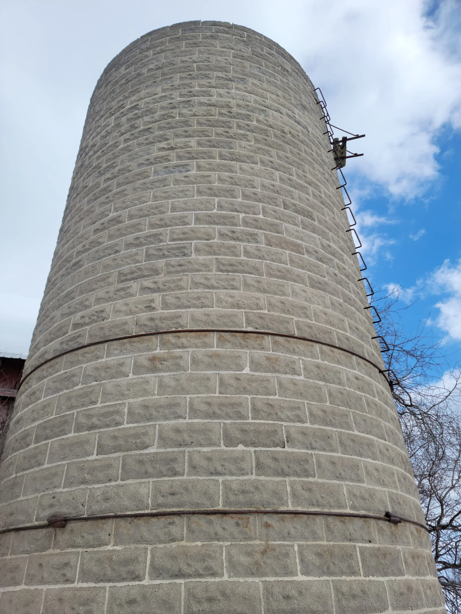 Looking straight up a concrete stave silo bound with steel hoops, a rusted ladder climbing its side against a blue sky. The worn blocks show its age.