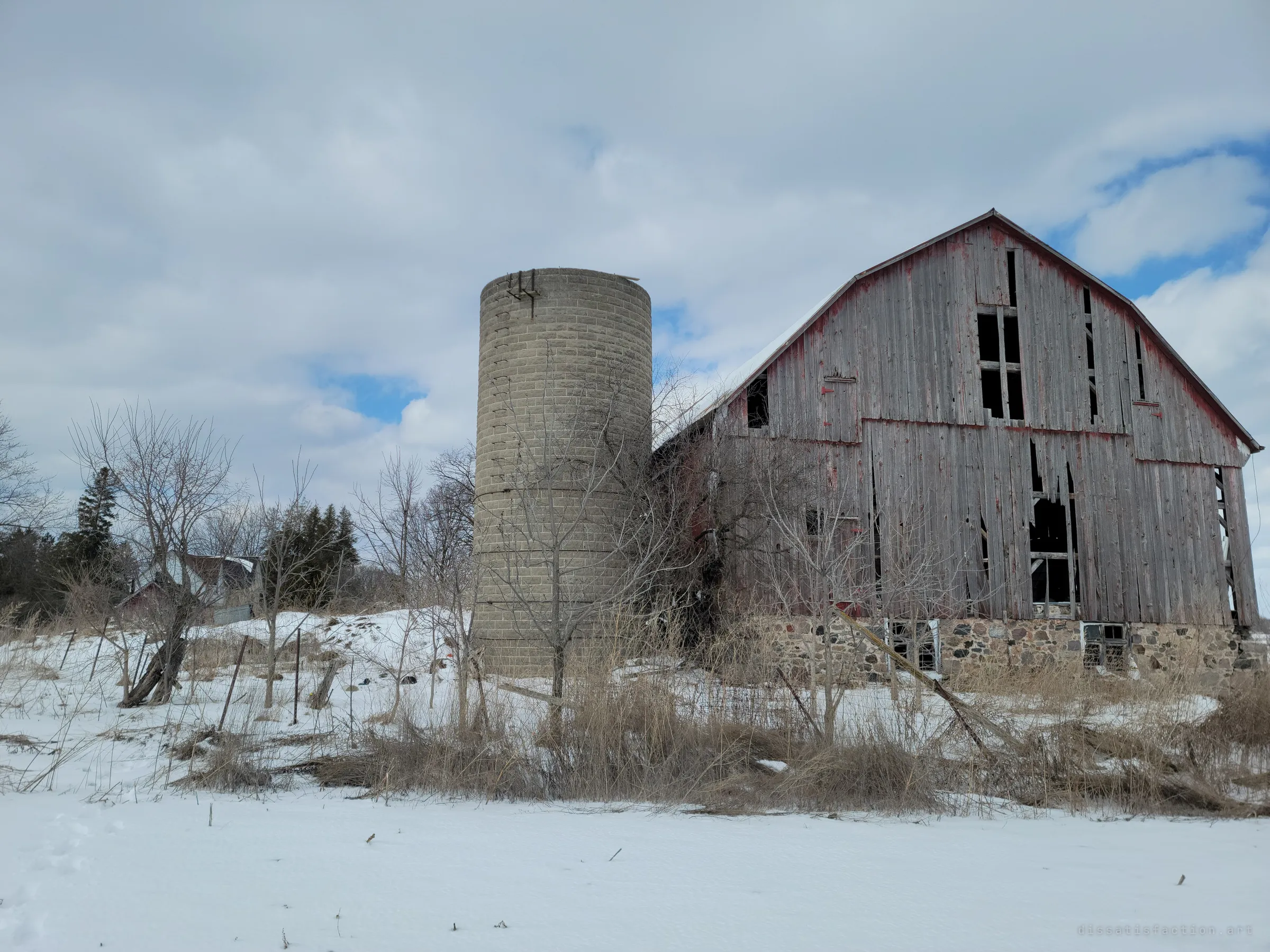 The weathered grey gambrel barn and its concrete silo stand above a snowy field, walls sagging and boards missing. Seen from outside, the whole farmstead looks like it is slowly giving way.